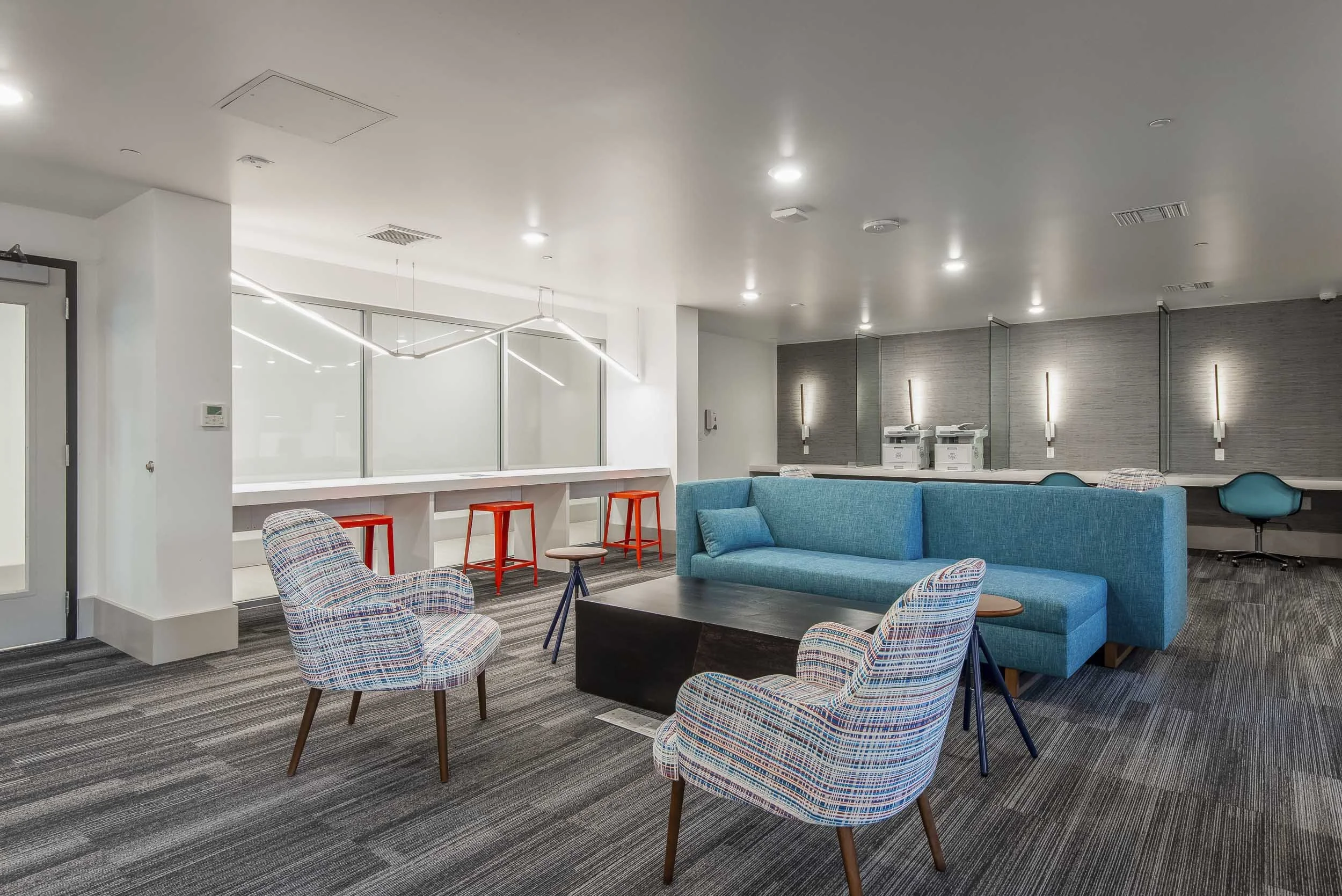 Modern lounge area with colorful patterned armchairs, a bright blue sofa, a black coffee table, and a row of red and white bar stools at a white counter. The space has gray striped carpeting, white walls, and contemporary lighting fixtures.