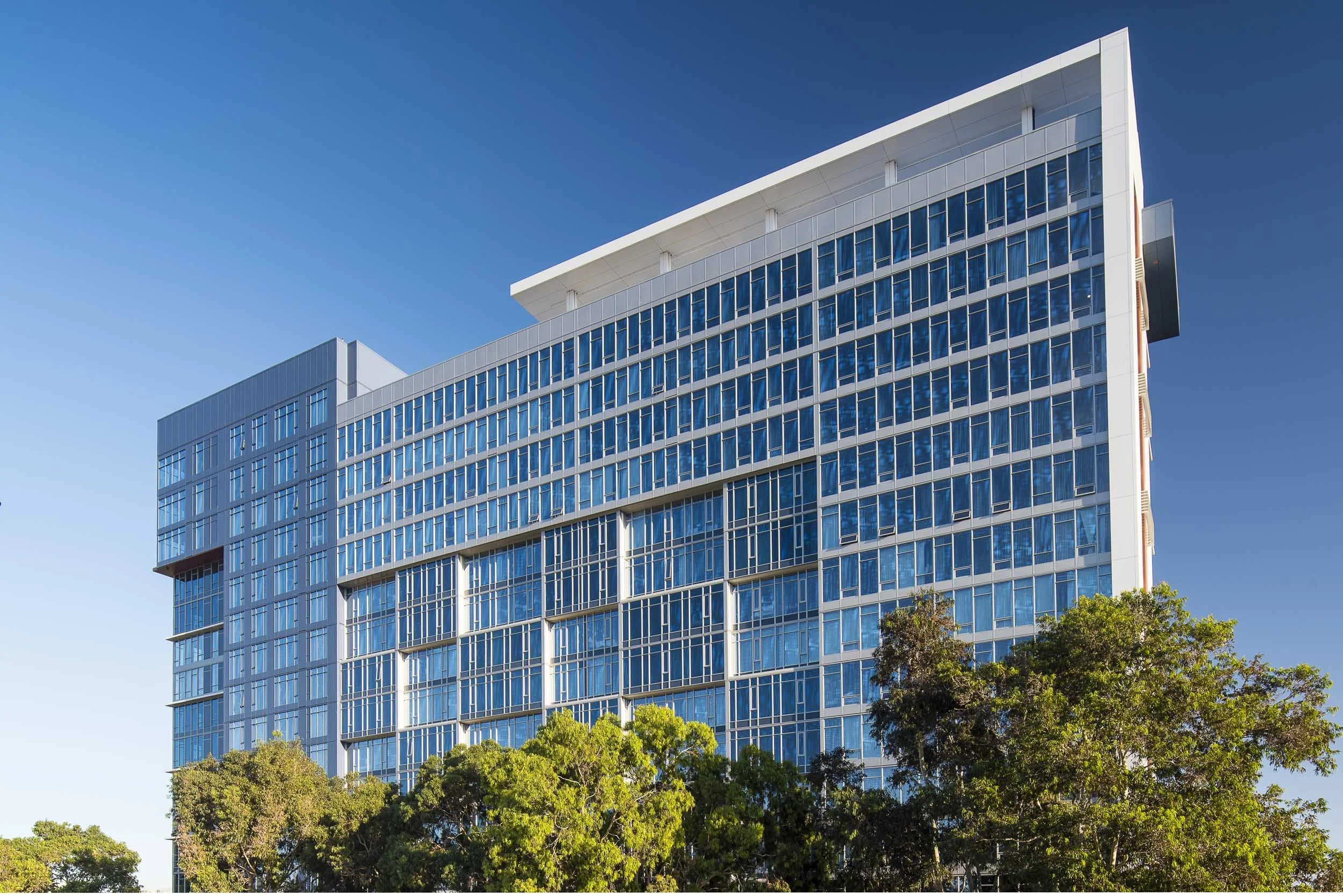 Modern glass high-rise building with multiple floors and reflective windows, surrounded by green trees against a clear blue sky.