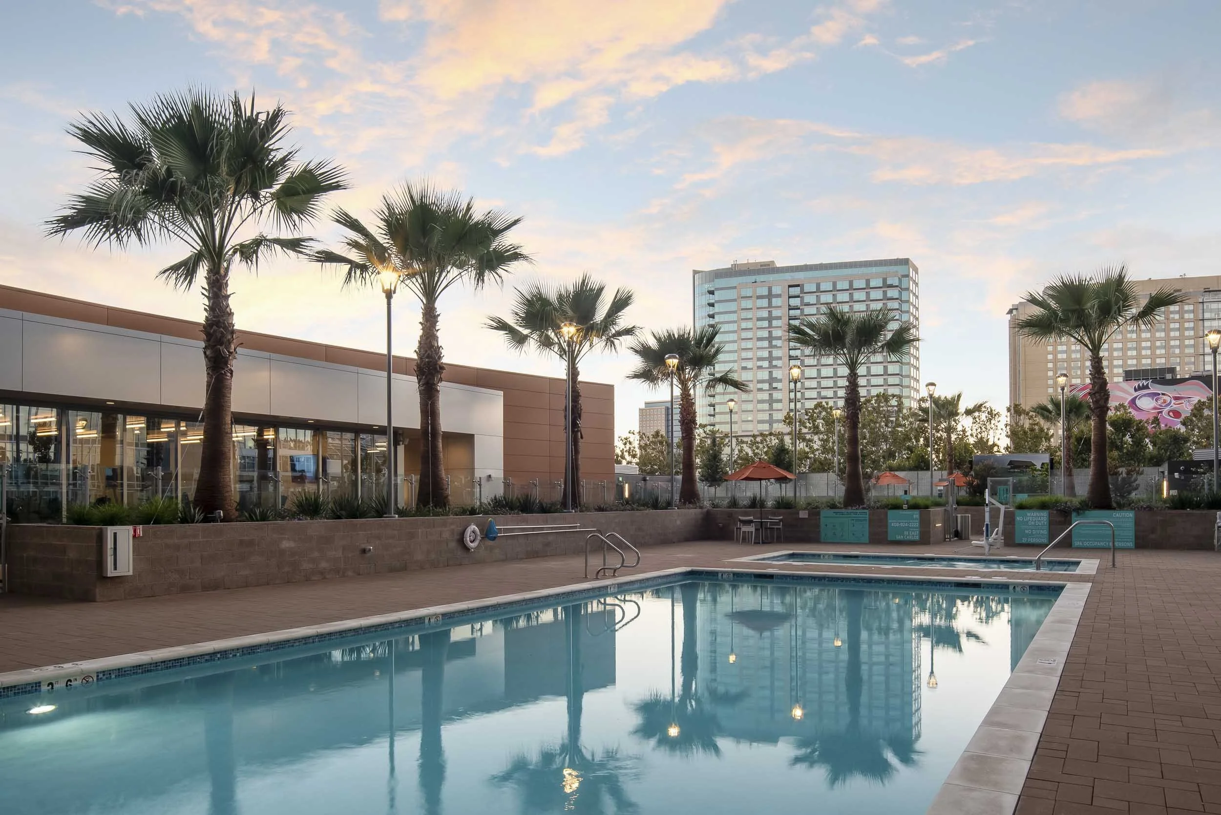 Empty outdoor swimming pool with palm trees, tall buildings in the background, and a partly cloudy sky at sunset.