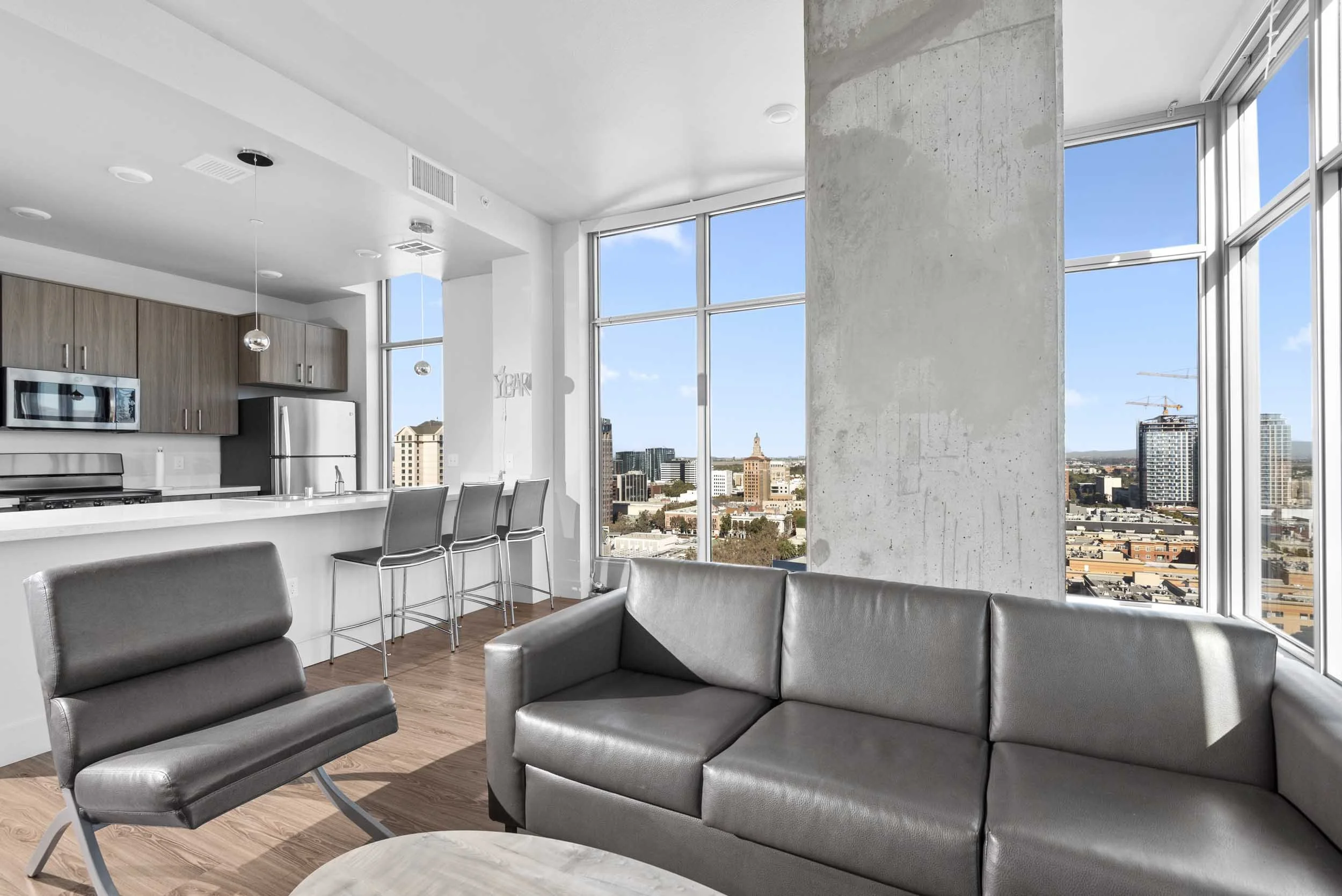 Modern open-concept living space with large windows showing city skyline, featuring gray couches, a kitchen with wooden cabinets, and a breakfast bar with chairs.