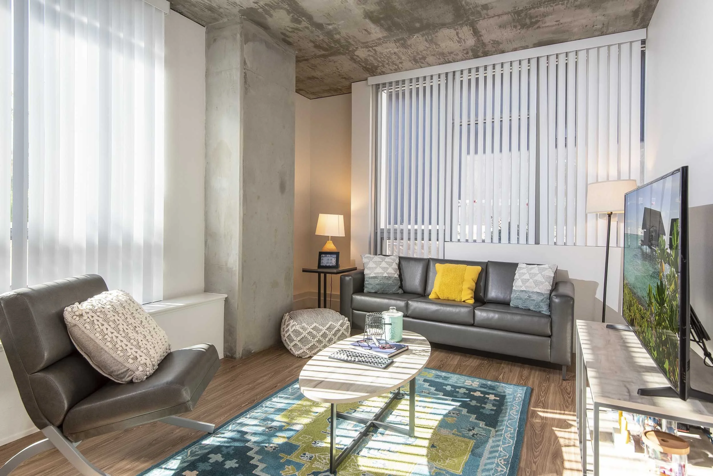 Modern living room with large windows covered by vertical blinds, concrete ceiling, gray sofa with yellow and patterned pillows, armchair, round coffee table, and a colorful area rug.