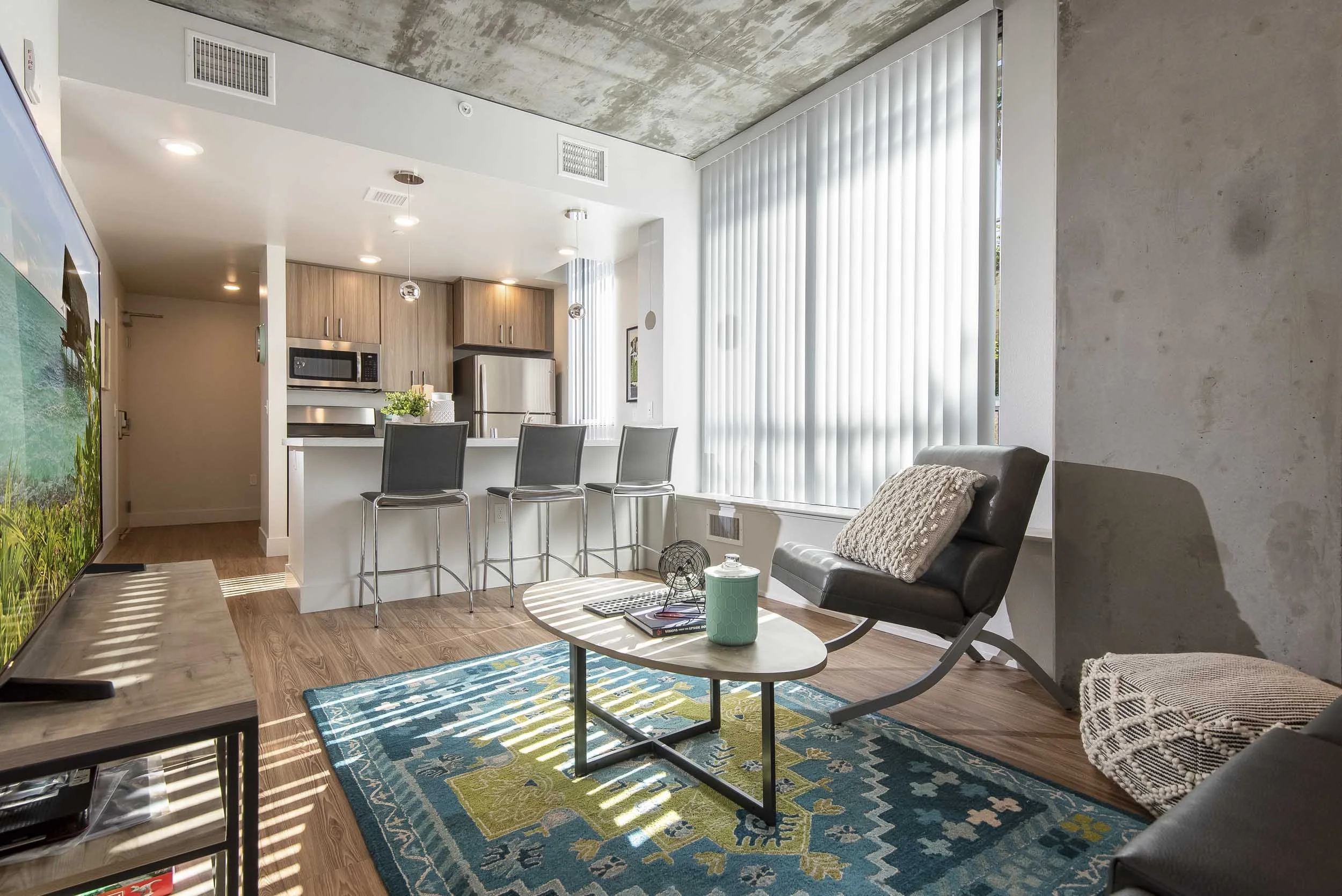 Modern living room with a black armchair, patterned blanket, and coffee table with teal tumbler, situated on colorful area rug. Kitchen in background with wooden cabinets, stainless steel appliances, and bar stools by a white counter. Large window with vertical blinds allowing sunlight, concrete ceiling, and wooden flooring.