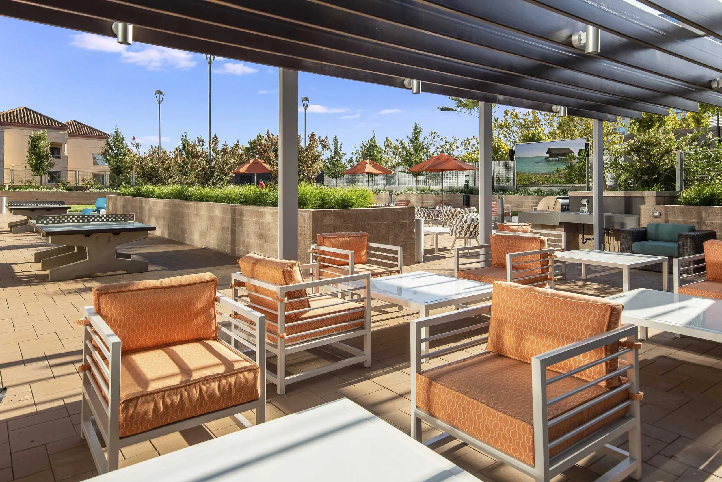 An outdoor patio area with orange cushioned chairs and tables, shaded by a dark wooden pergola, featuring a view of trees, umbrellas, and a large screen displaying a tropical scene.