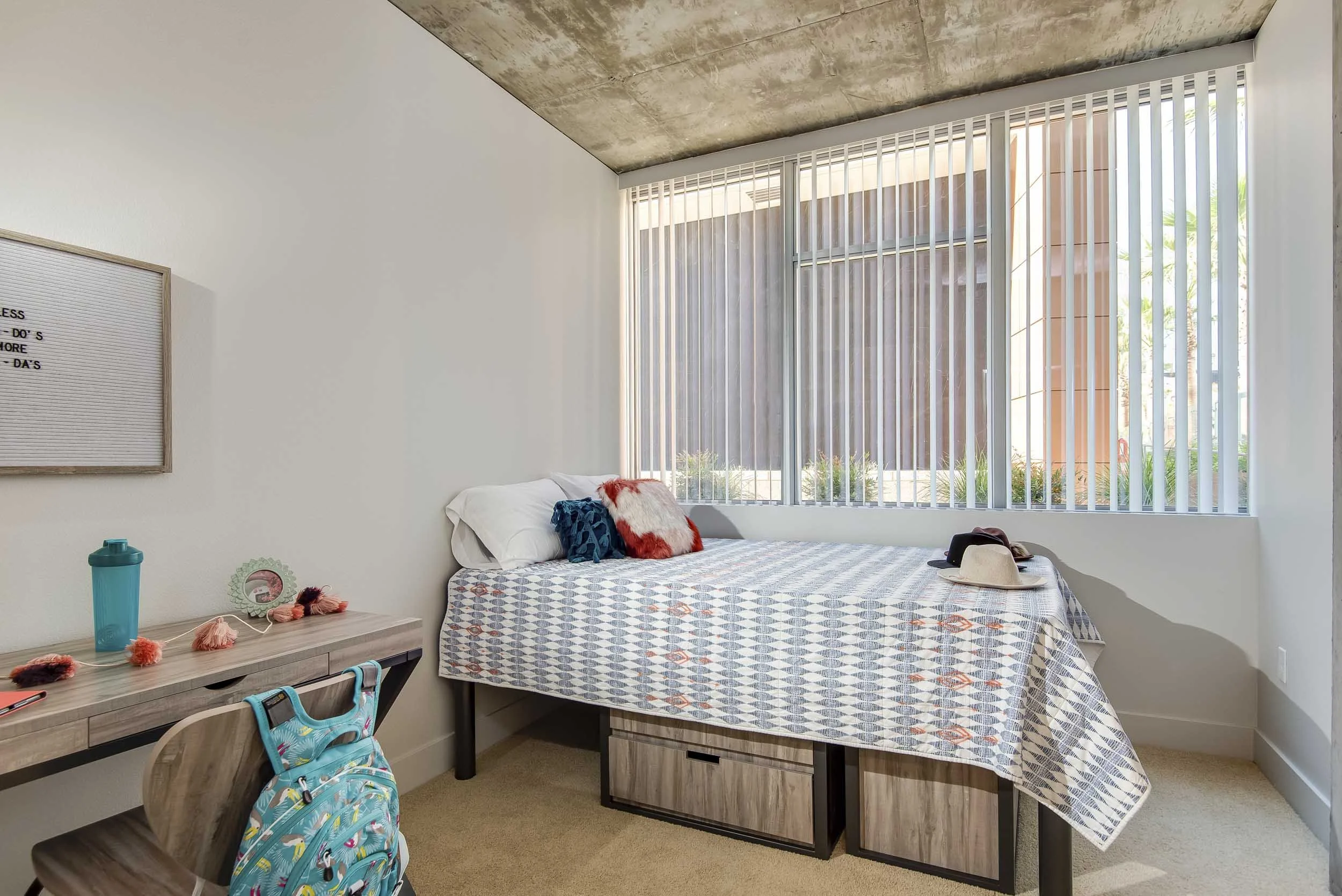 A bedroom with a bed near a large window with vertical blinds. There are pillows and a blanket on the bed, and a hat and backpack on the edge. A side table has a water bottle, a mirror, and tassel string decorations.