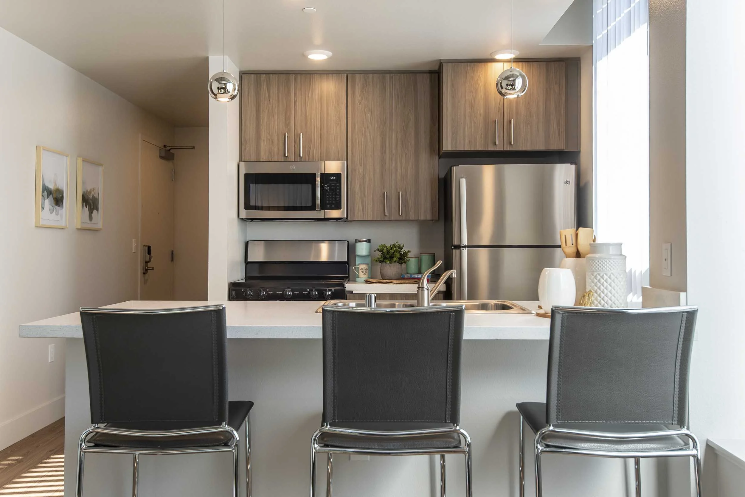Modern kitchen with wooden cabinets, stainless steel refrigerator, microwave, black stove, and white kitchen island with three black chairs. Decor includes a potted plant, vases, and art on the walls. Large window with vertical blinds lets in natural light.