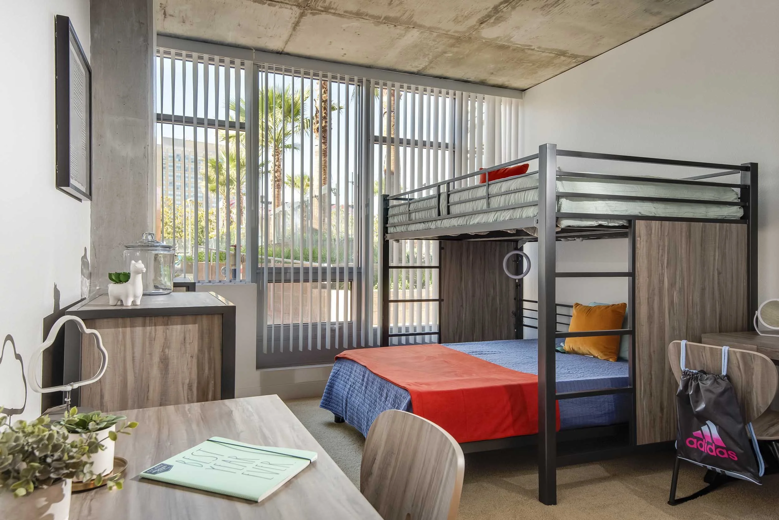 A modern apartment room featuring a bunk bed with a desk underneath, large windows with vertical blinds showing palm trees outside, a wooden dining table with green plants, and various decorative items.