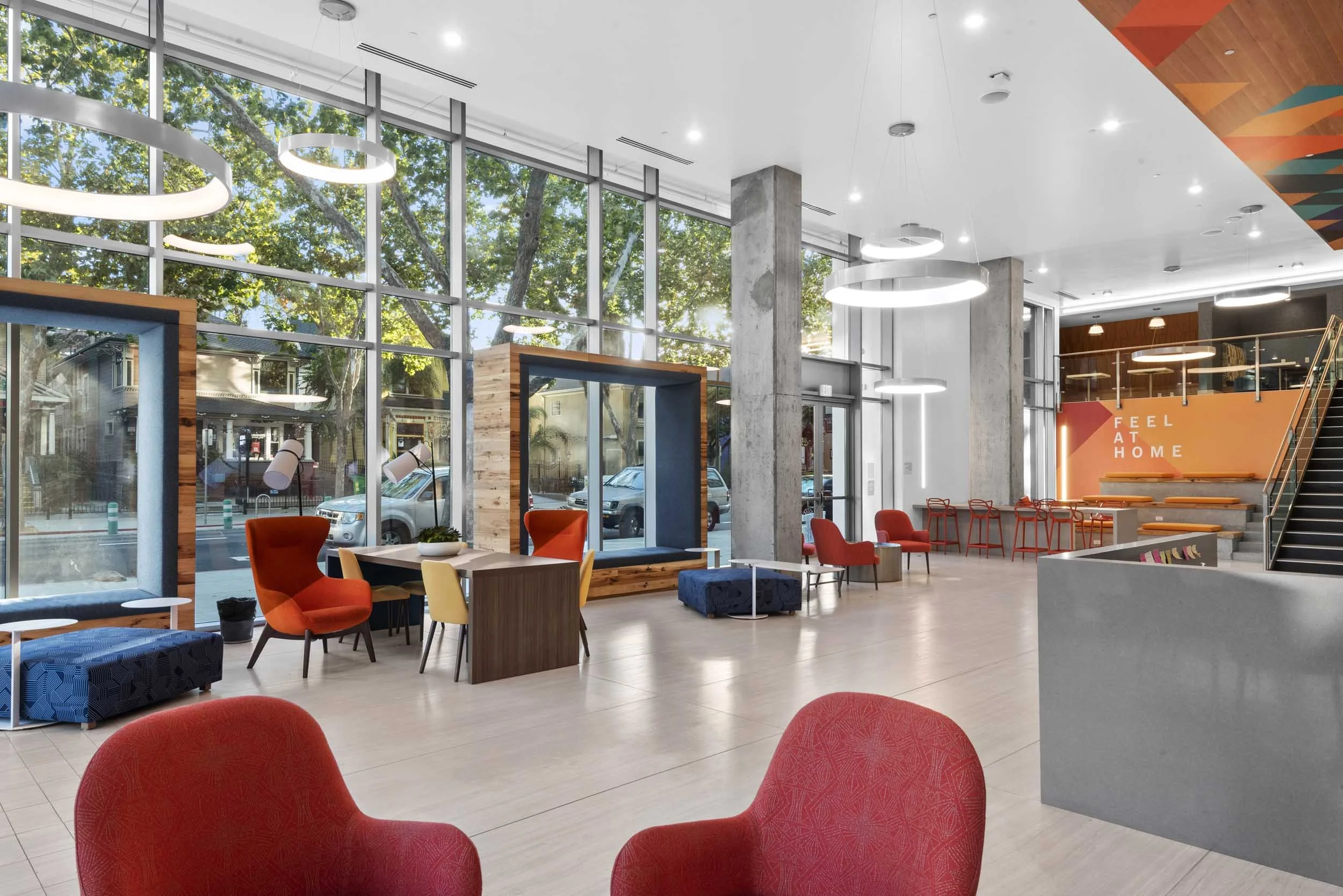 Modern indoor space with large glass windows showing trees outside, featuring various colorful chairs, tables, and decorative elements, with a staircase and a mural that says "Feel at Home."