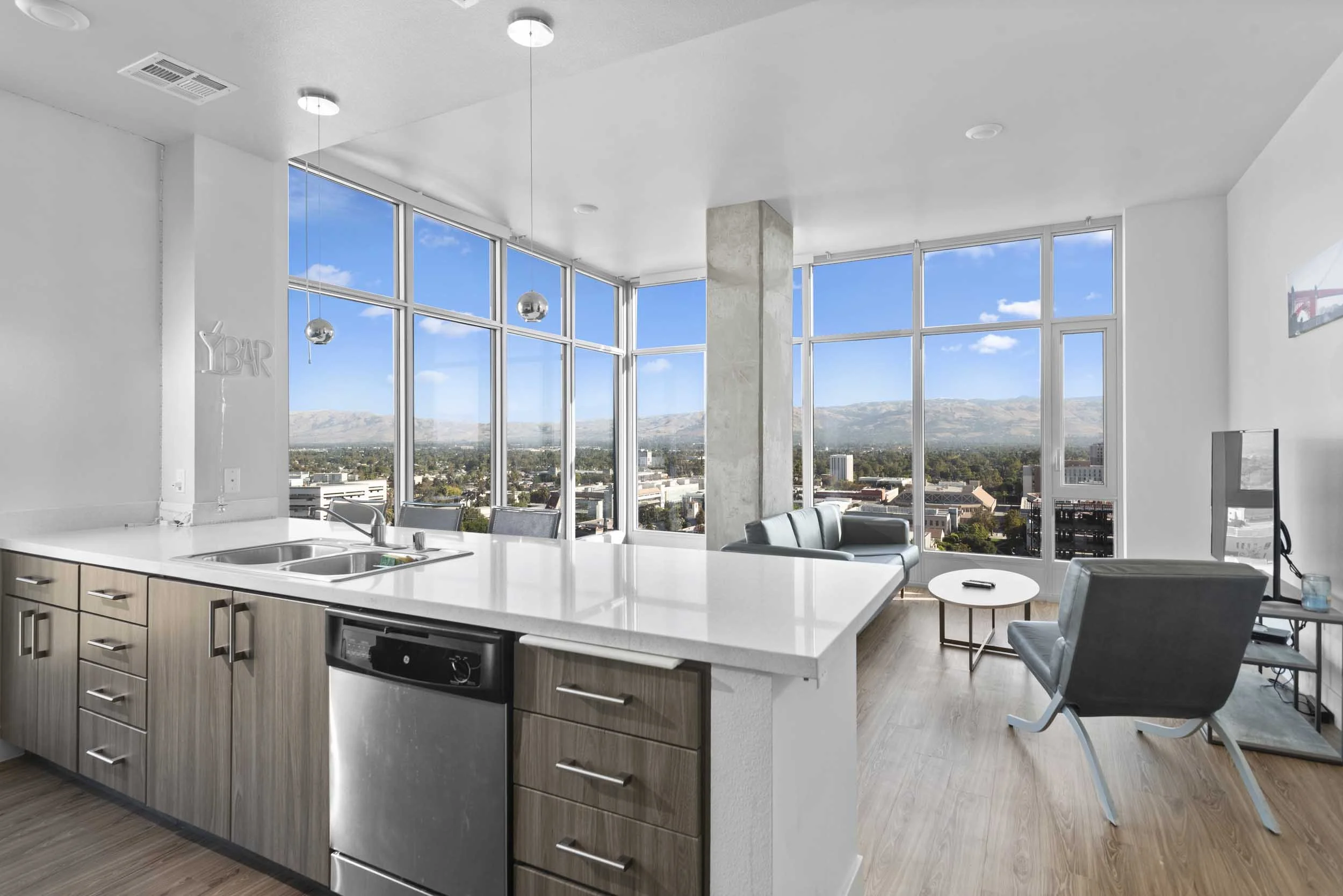 Modern apartment kitchen with large windows showing city and mountain view, kitchen island with sink and dishwasher, and living area with chairs and TV.
