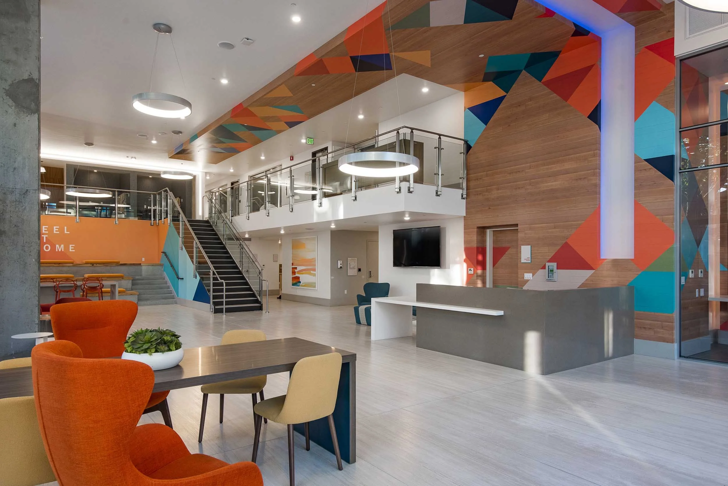Modern indoor space with colorful chairs, a table with a plant, a staircase, a wall with geometric art, and a large TV.