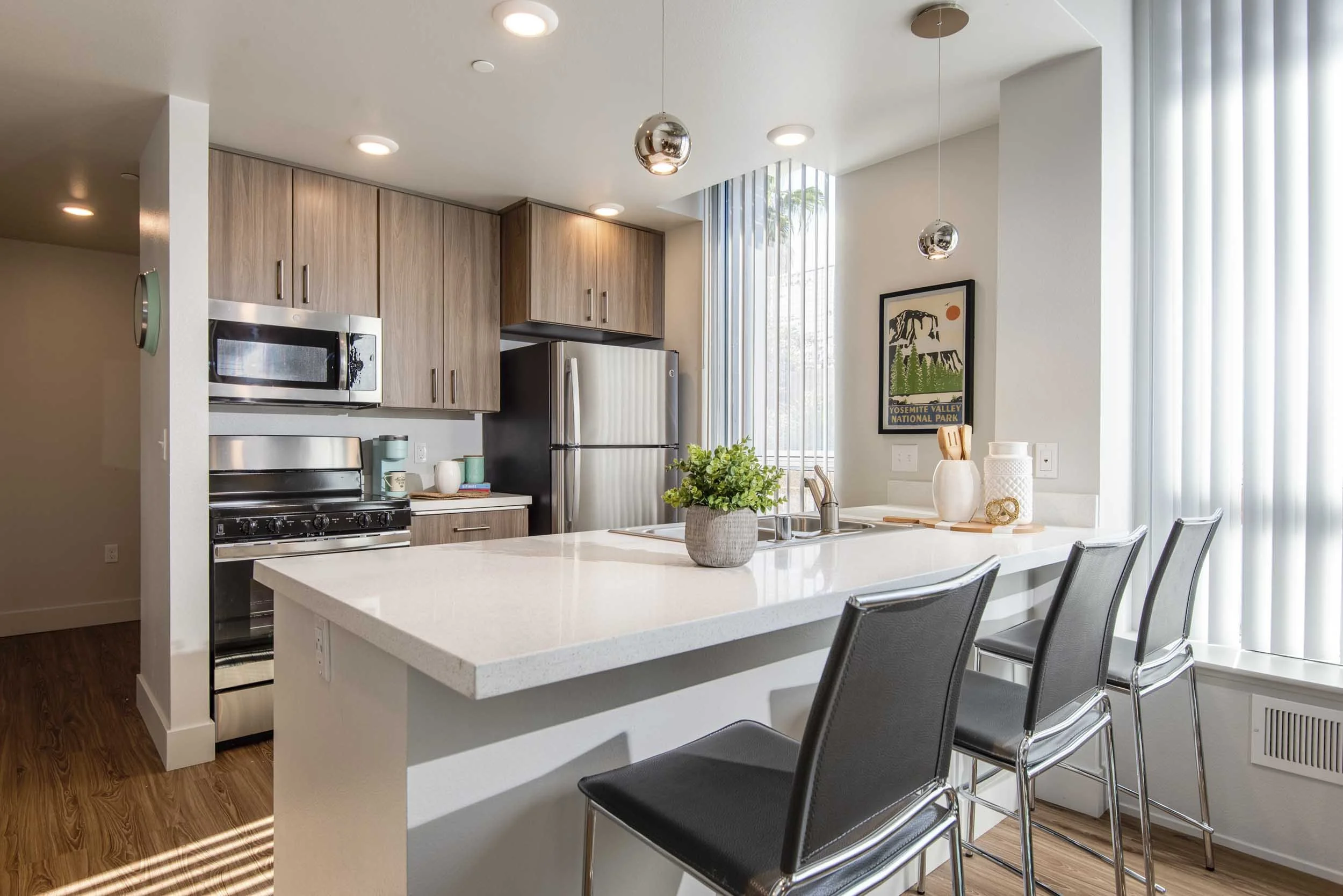 Modern kitchen with a white island counter, black chairs, wooden cabinetry, stainless steel appliances, potted plant, and artwork close to large windows with vertical blinds.