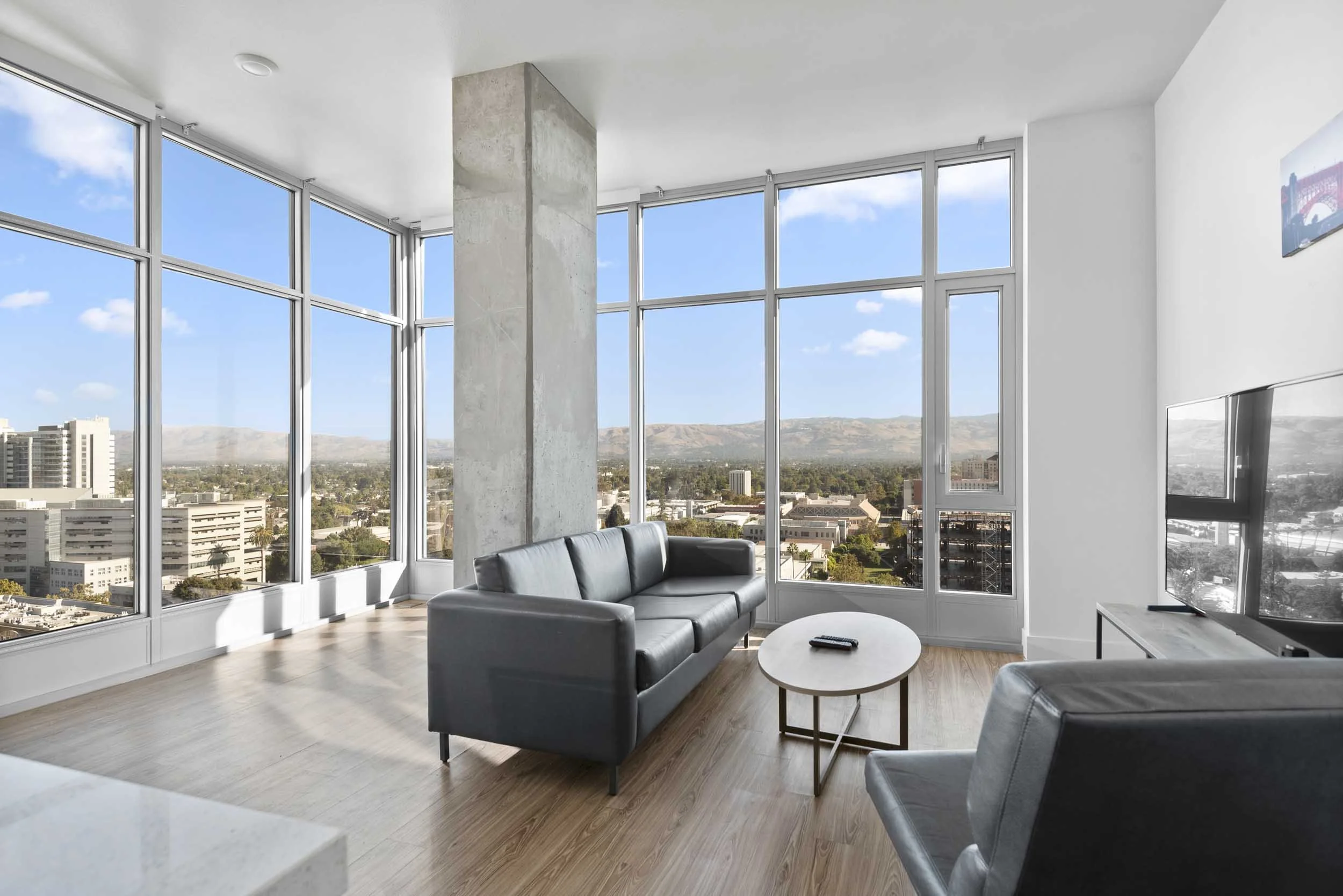Interior of a modern high-rise apartment with large floor-to-ceiling windows showing cityscape and mountains in the distance. The room features a gray sofa, a small round white coffee table with a remote on it, and a television on the wall.