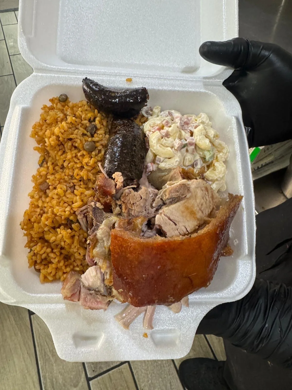A foam takeout container filled with rice, stuffed grape leaves, potato salad, roasted pork, and a piece of roasted pork shoulder.