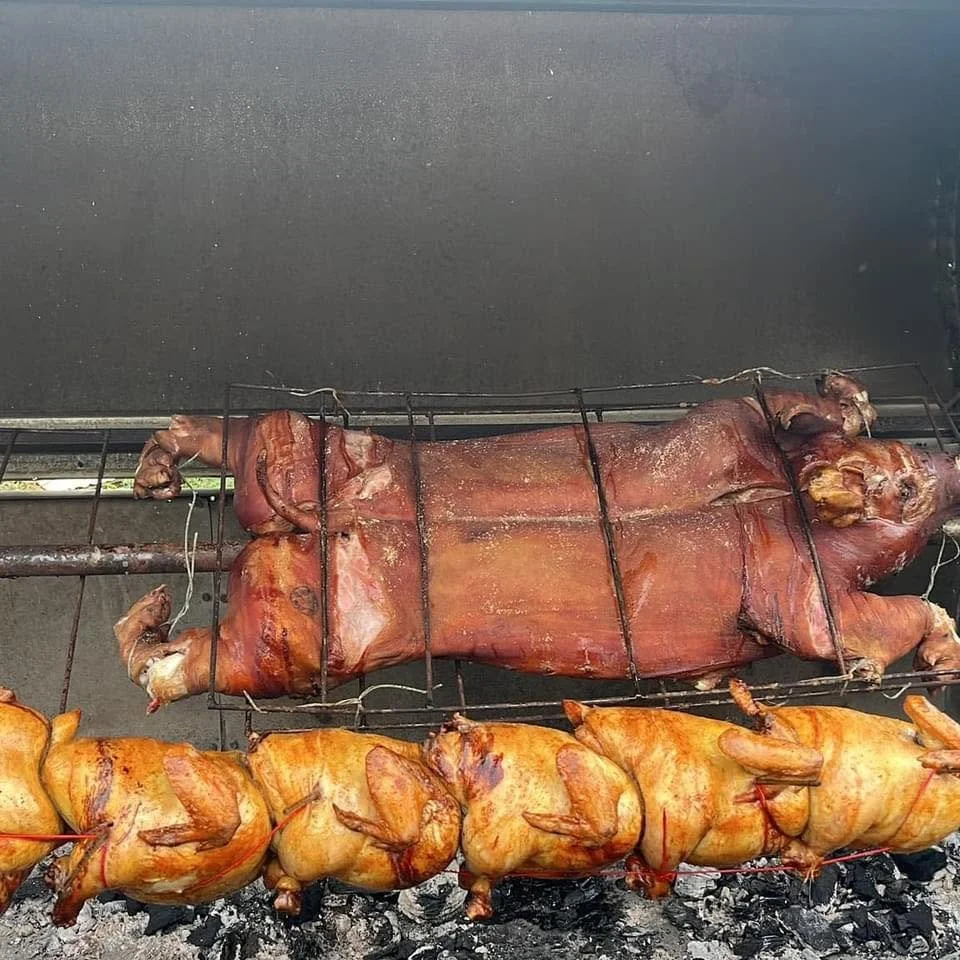 Whole pig roasting on a spit over charcoal grill, with skewered chicken below it.