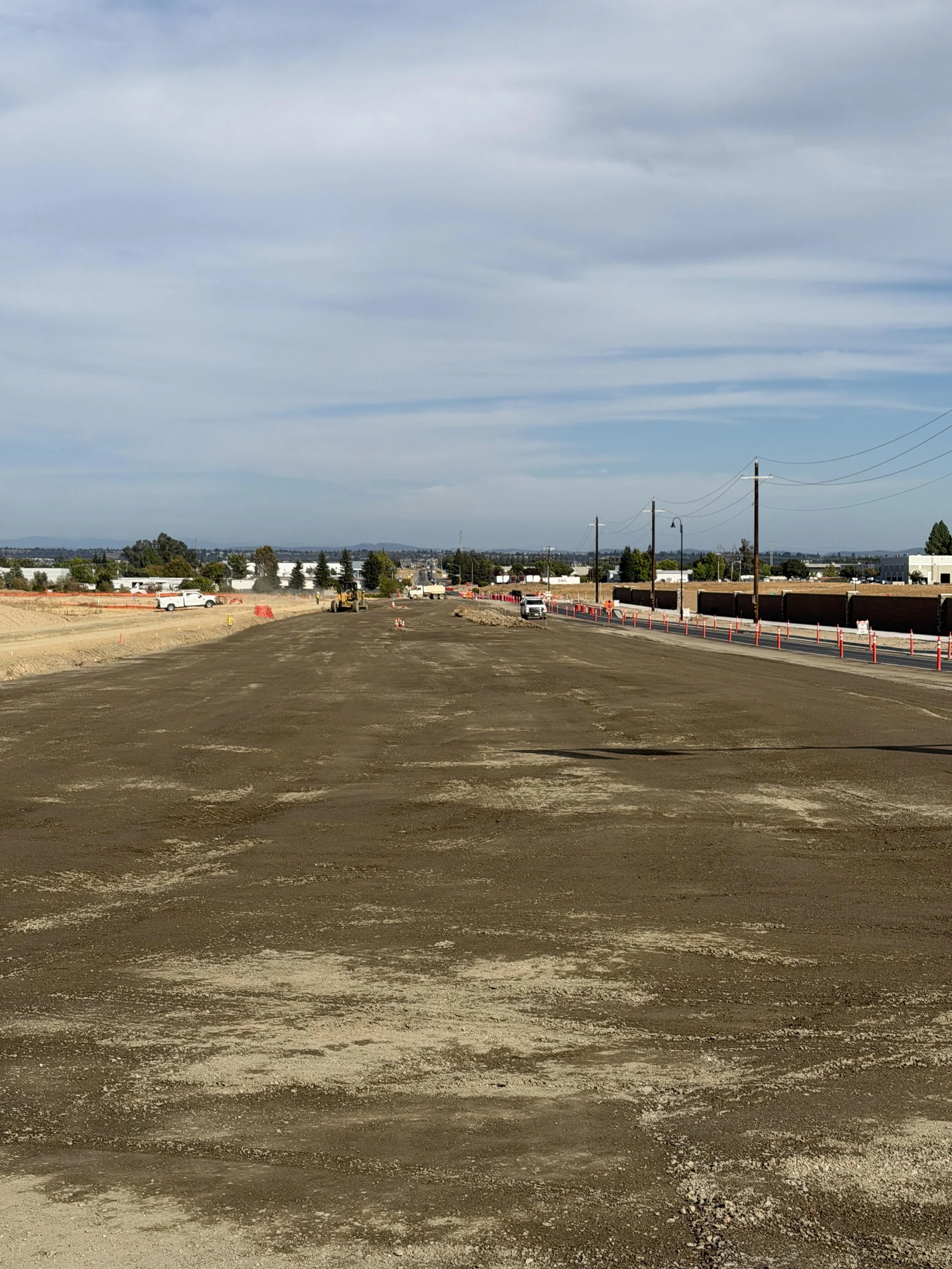 Road under construction with vehicles, construction cones, and utility poles along the side. Overcast sky in the background.
