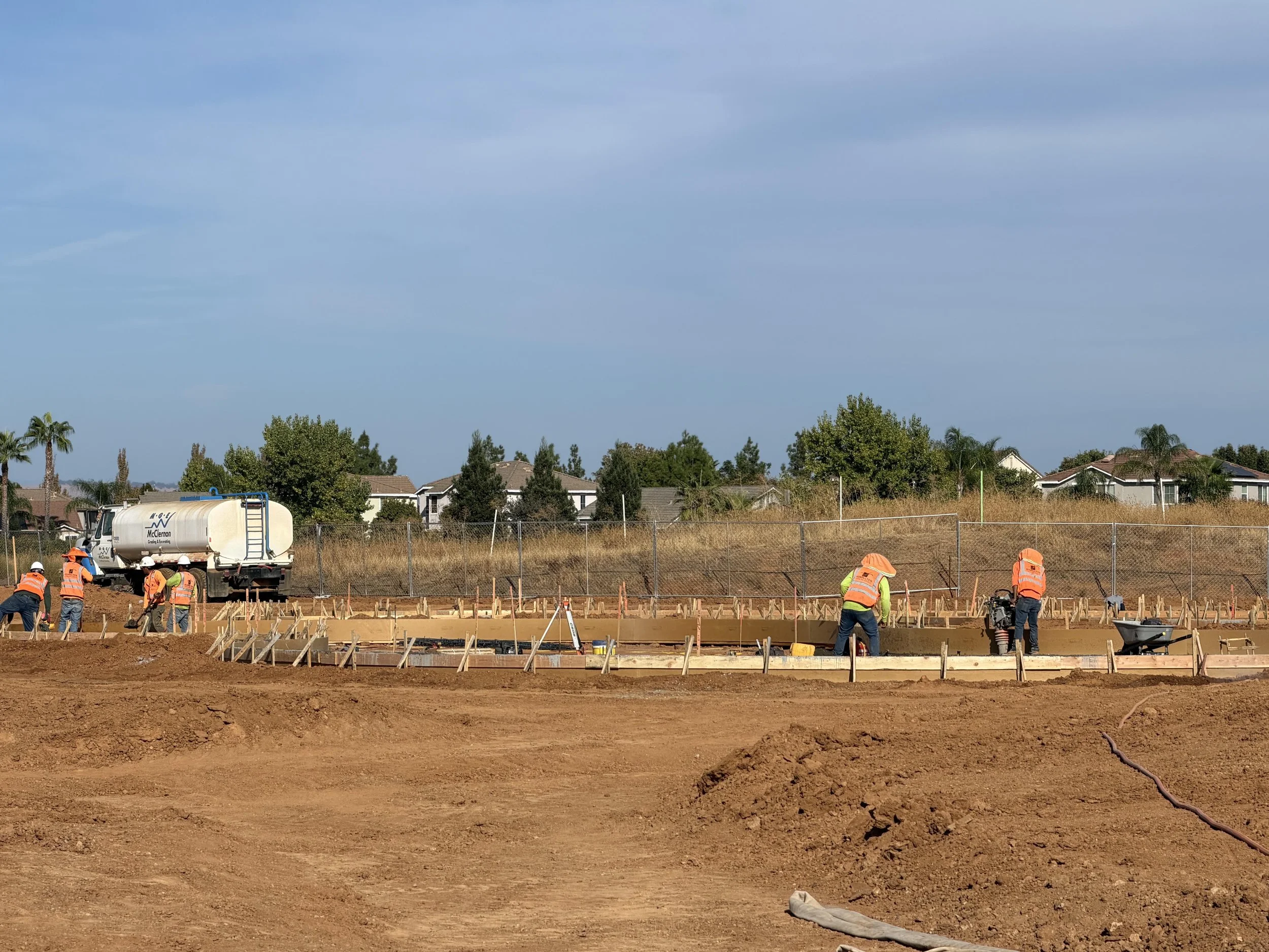 Construction workers in orange safety vests and helmets working on a building site with wooden forms for concrete foundation, a truck with a water tank, and residential houses in the background under a cloudy sky.