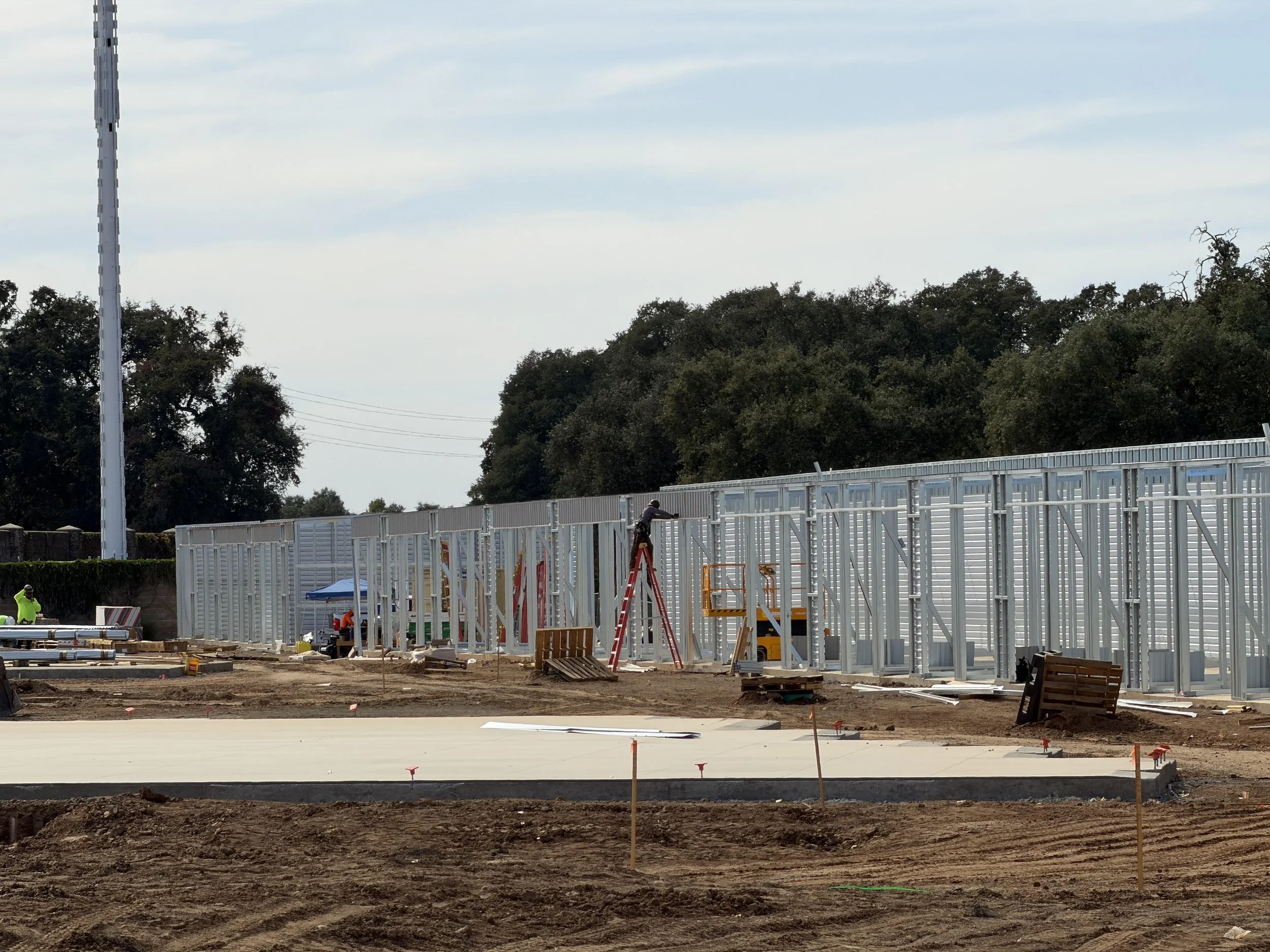Construction site with metal framing walls, construction workers, tools, and equipment in progress, surrounded by trees and an overcast sky.