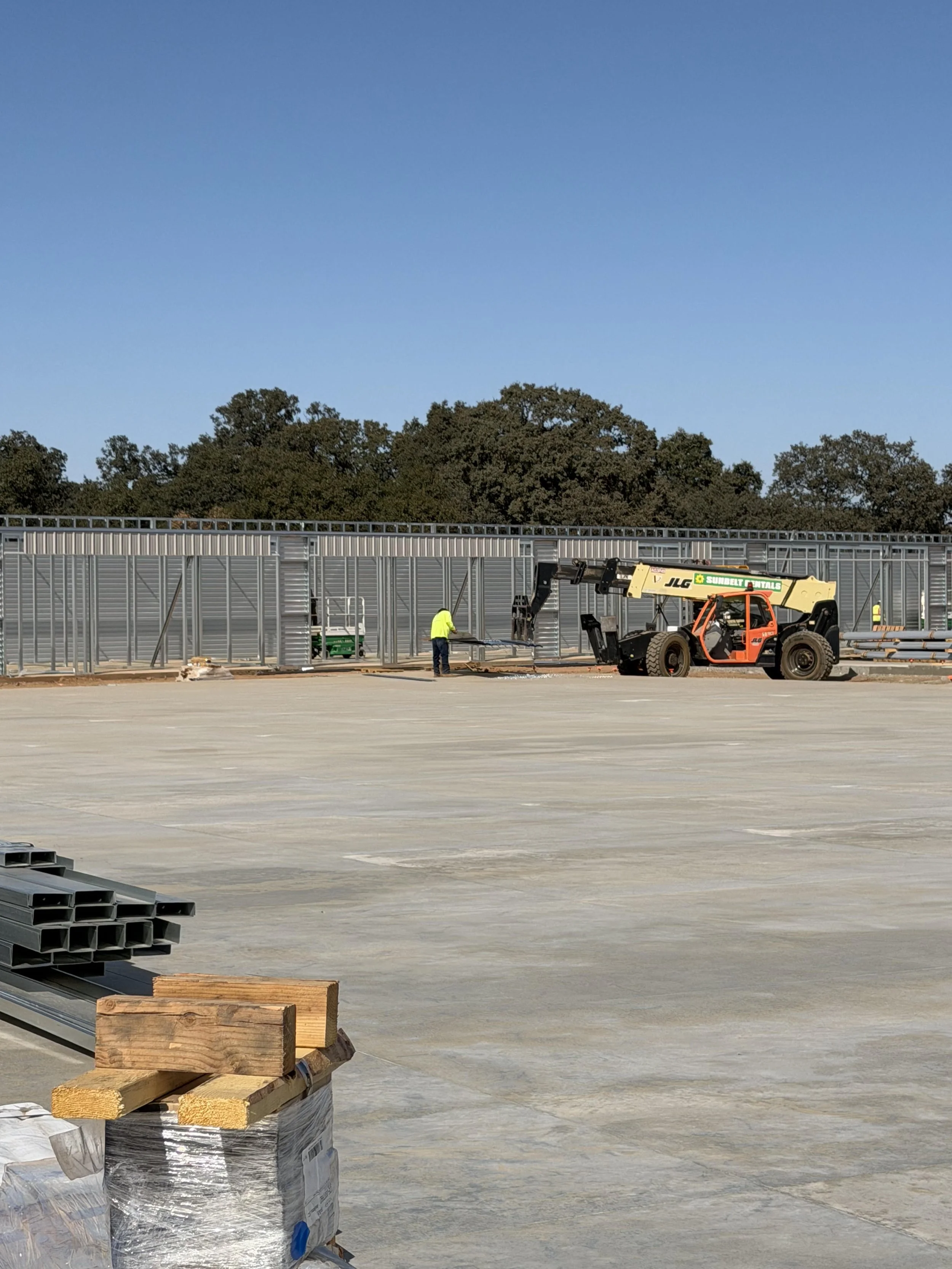Construction site with workers and heavy equipment, a metal wall, stacked pipes, and wooden beams on concrete ground under a clear blue sky.