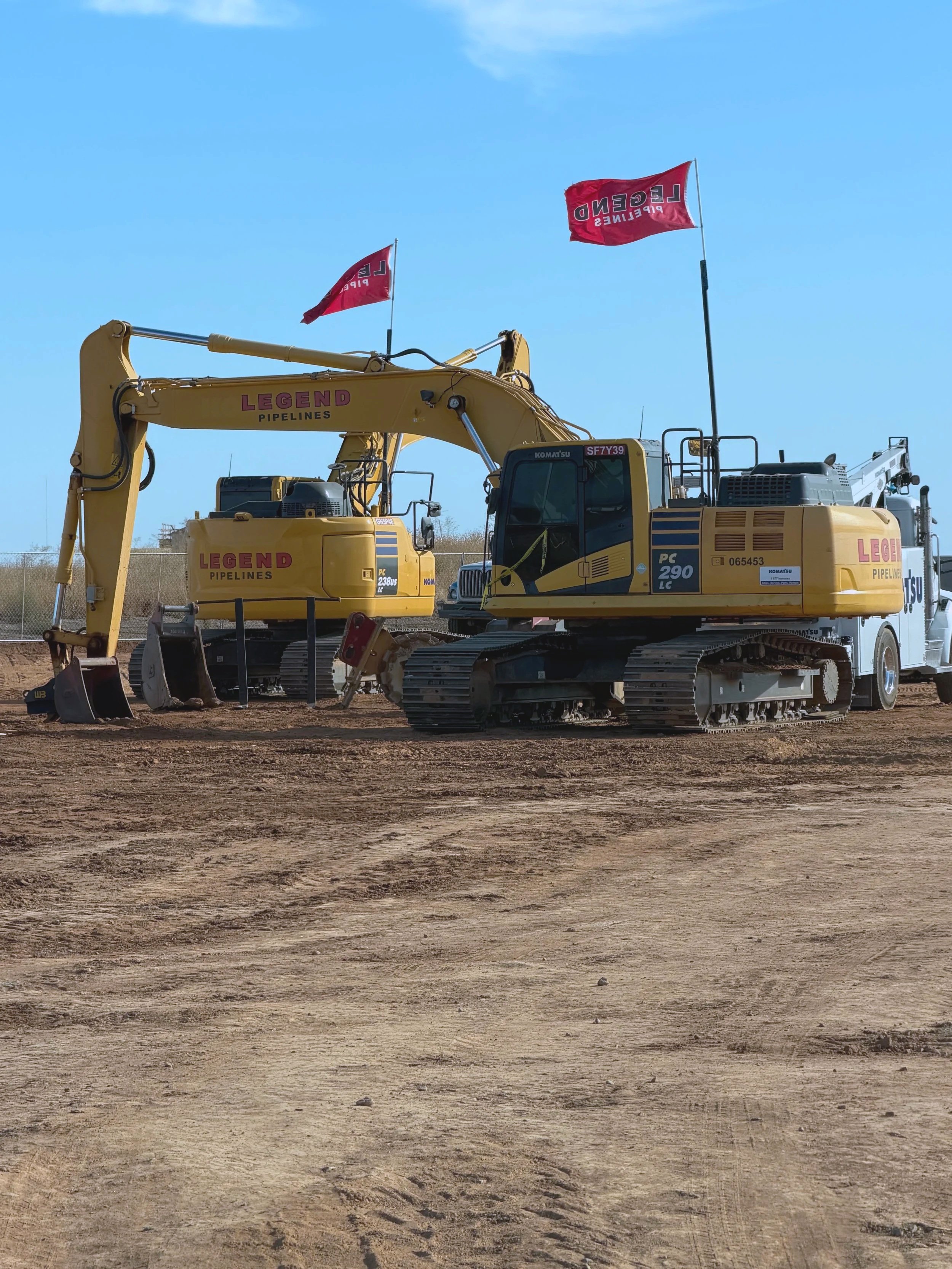 A construction site with two yellow excavators and two red flags that read "LEND PIPELINES." The ground is bare dirt, and the sky is clear with minimal clouds.