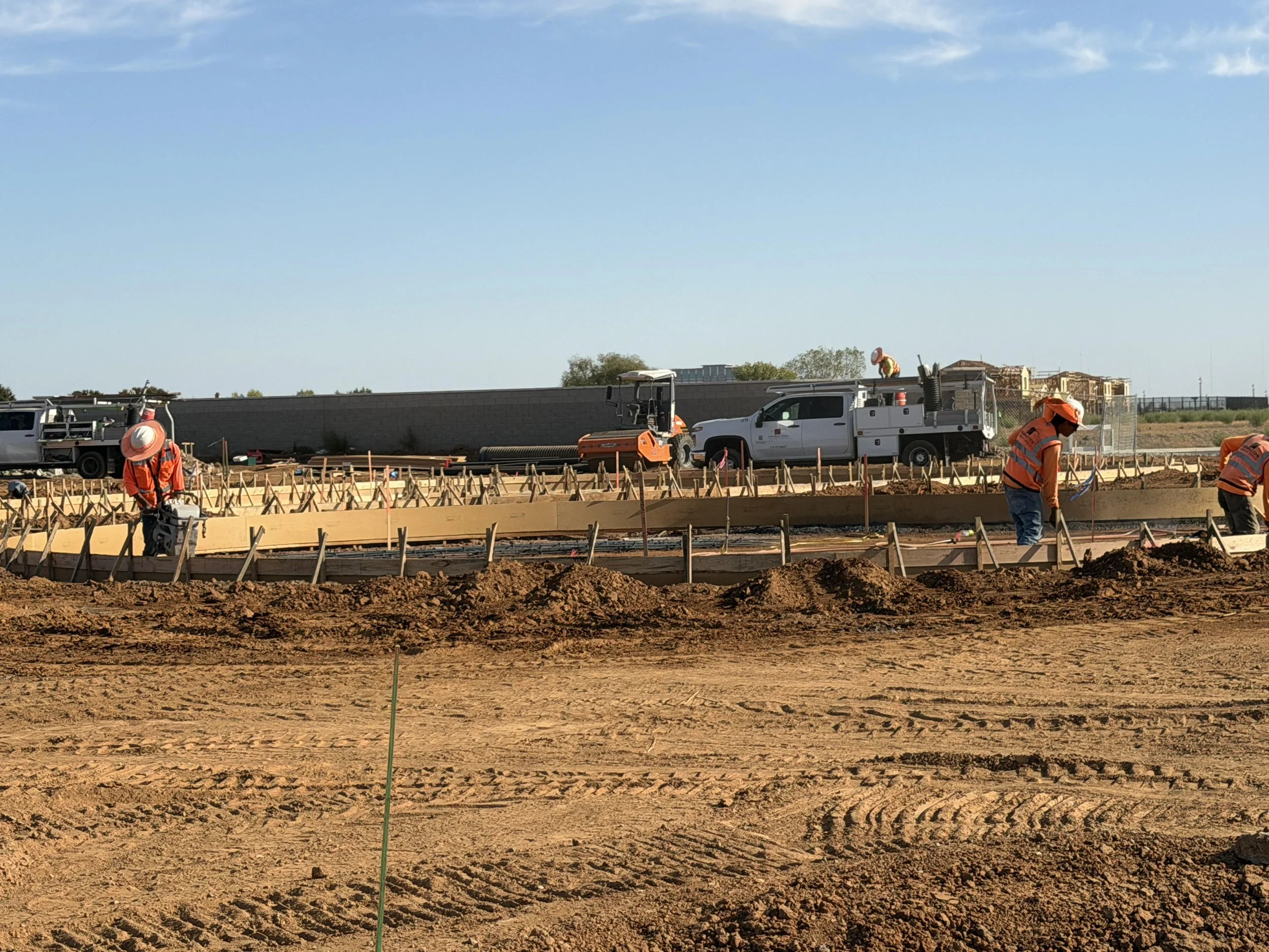 Construction workers in orange safety vests and helmets working on a building site with construction vehicles, wooden framework, and a high wall in the background under a clear blue sky.
