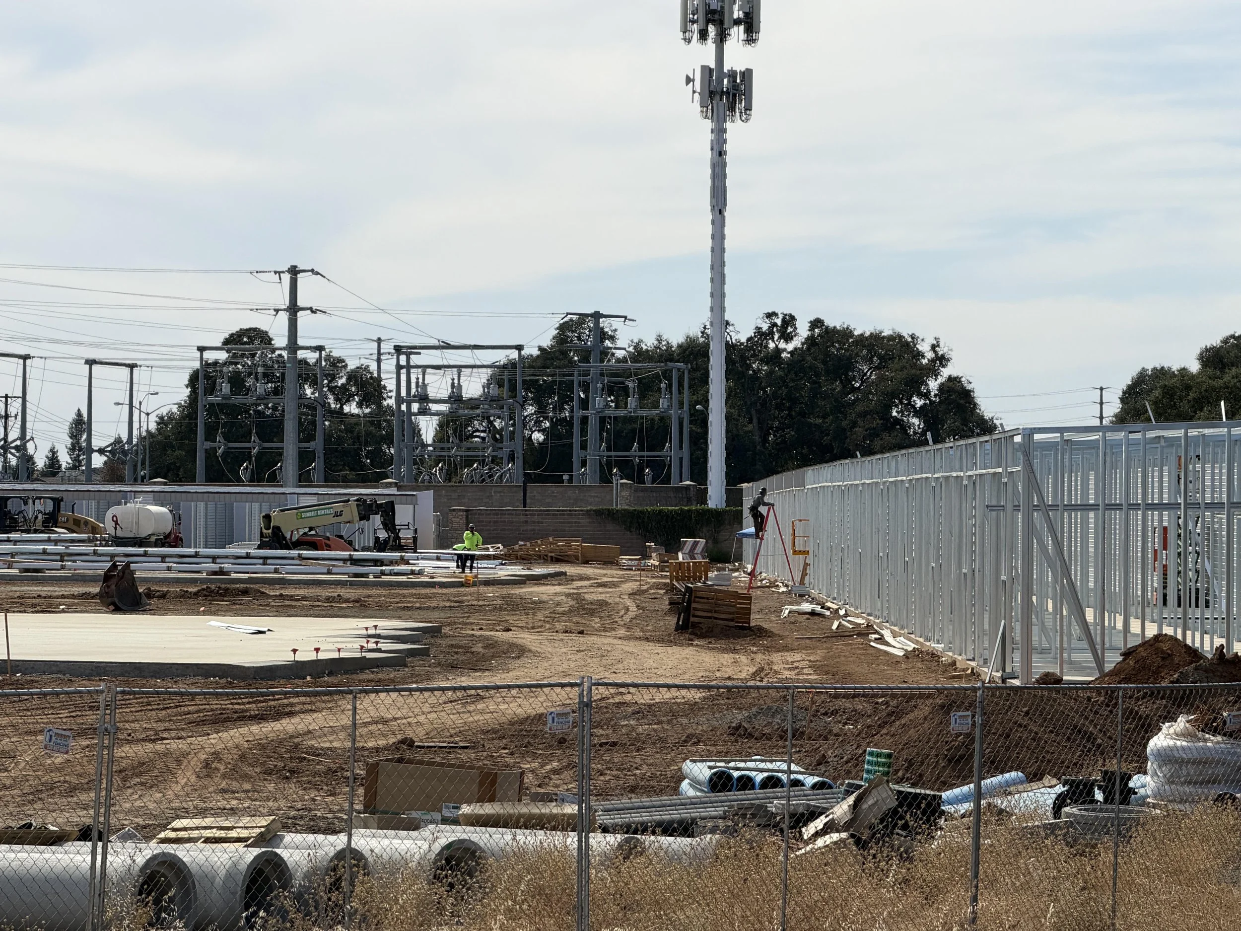 Construction site with electrical infrastructure, workers, machinery, and materials, fenced off with a chain-link fence.