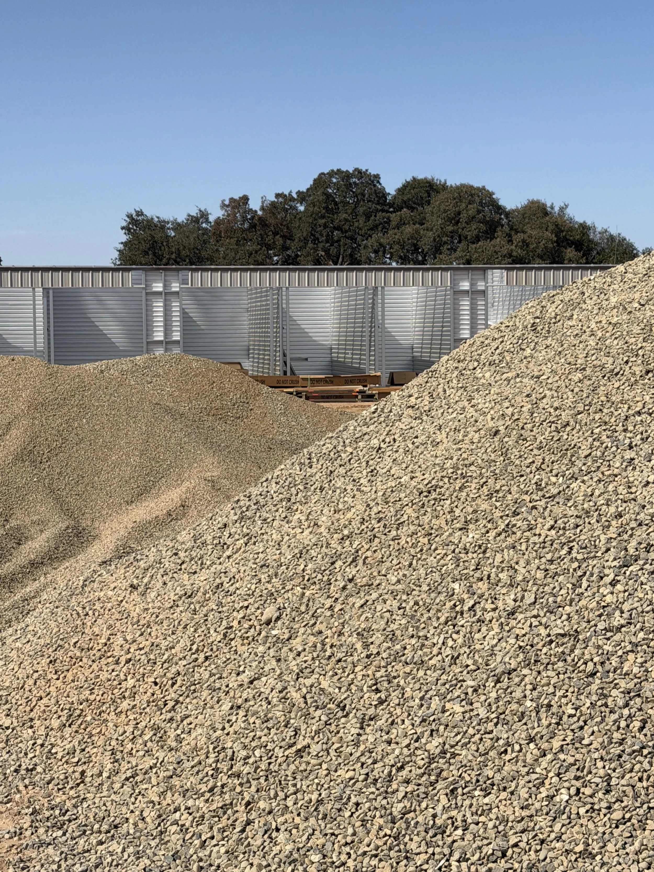 Large gravel piles in front of a metal construction barrier with trees in the background and a clear blue sky