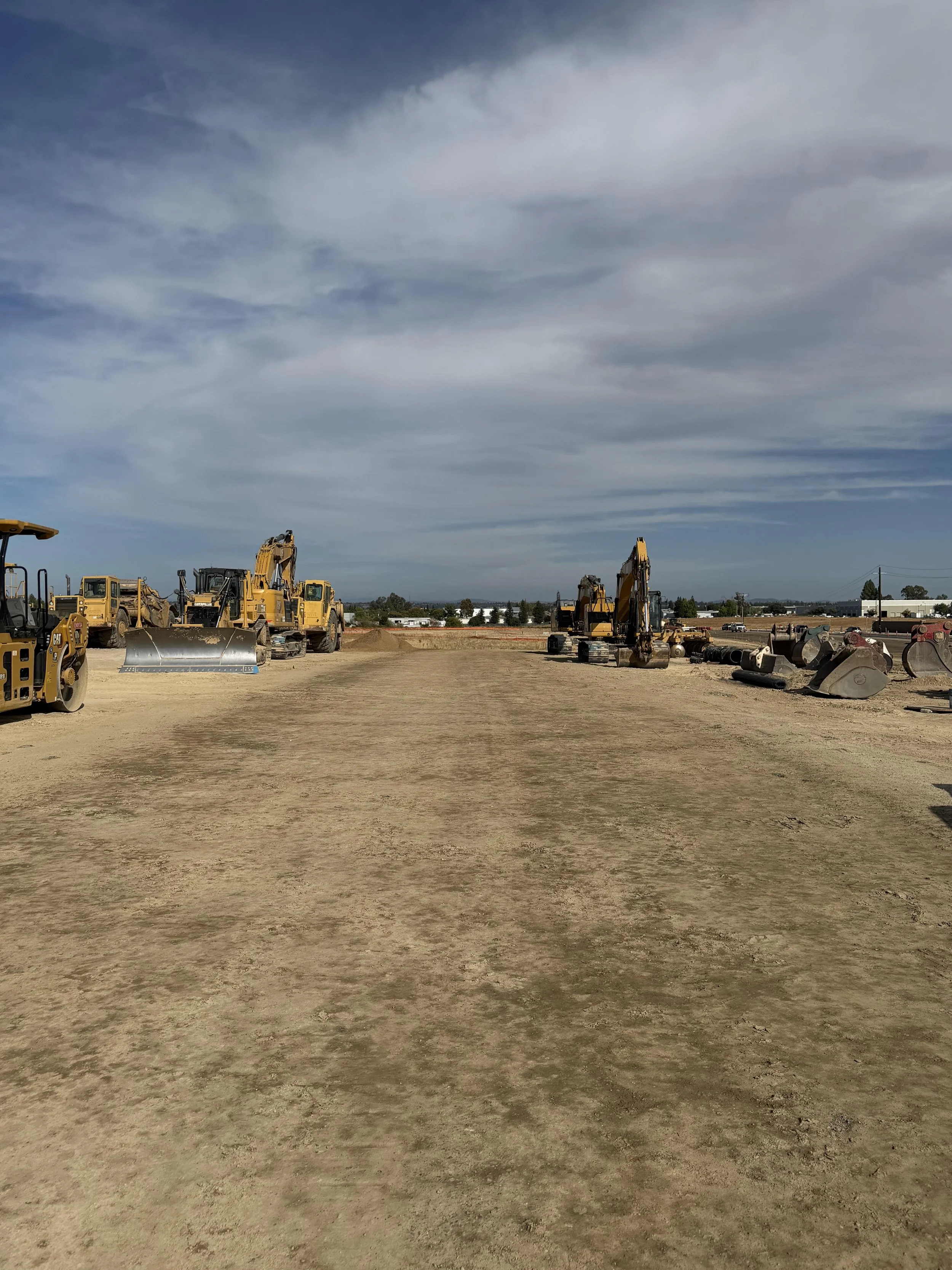 Construction site with yellow excavators and bulldozers on dirt ground, under a cloudy sky.