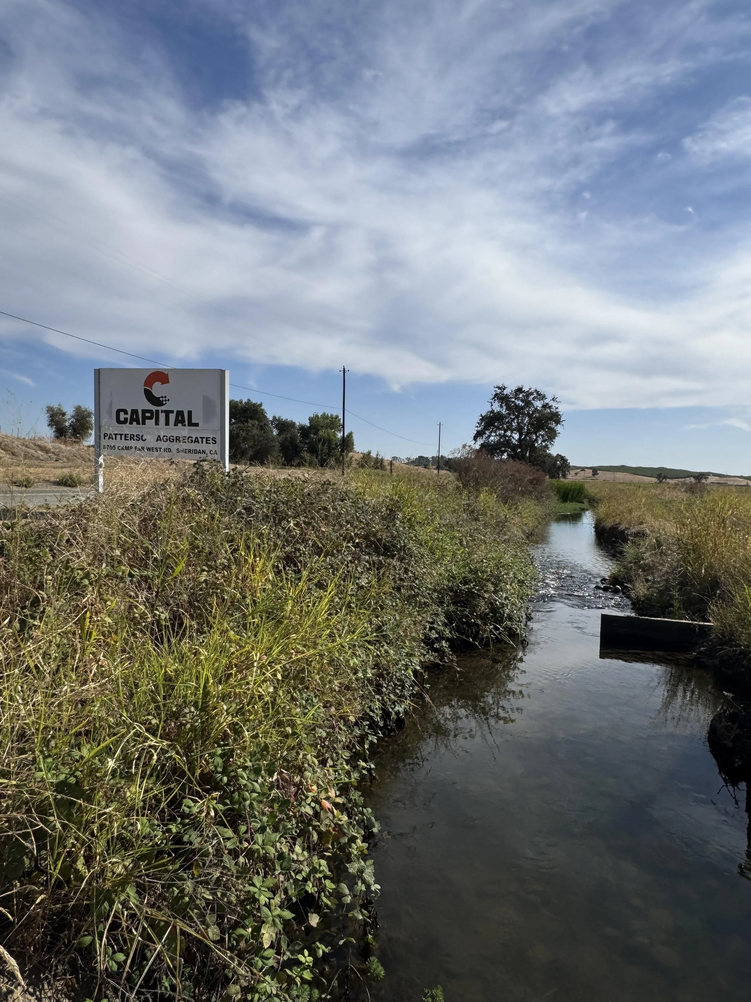 A small creek runs through a grassy area with bushes and trees. A sign for Capital Patterson Aggregates is positioned near the creek, with a blue sky and wispy clouds above.