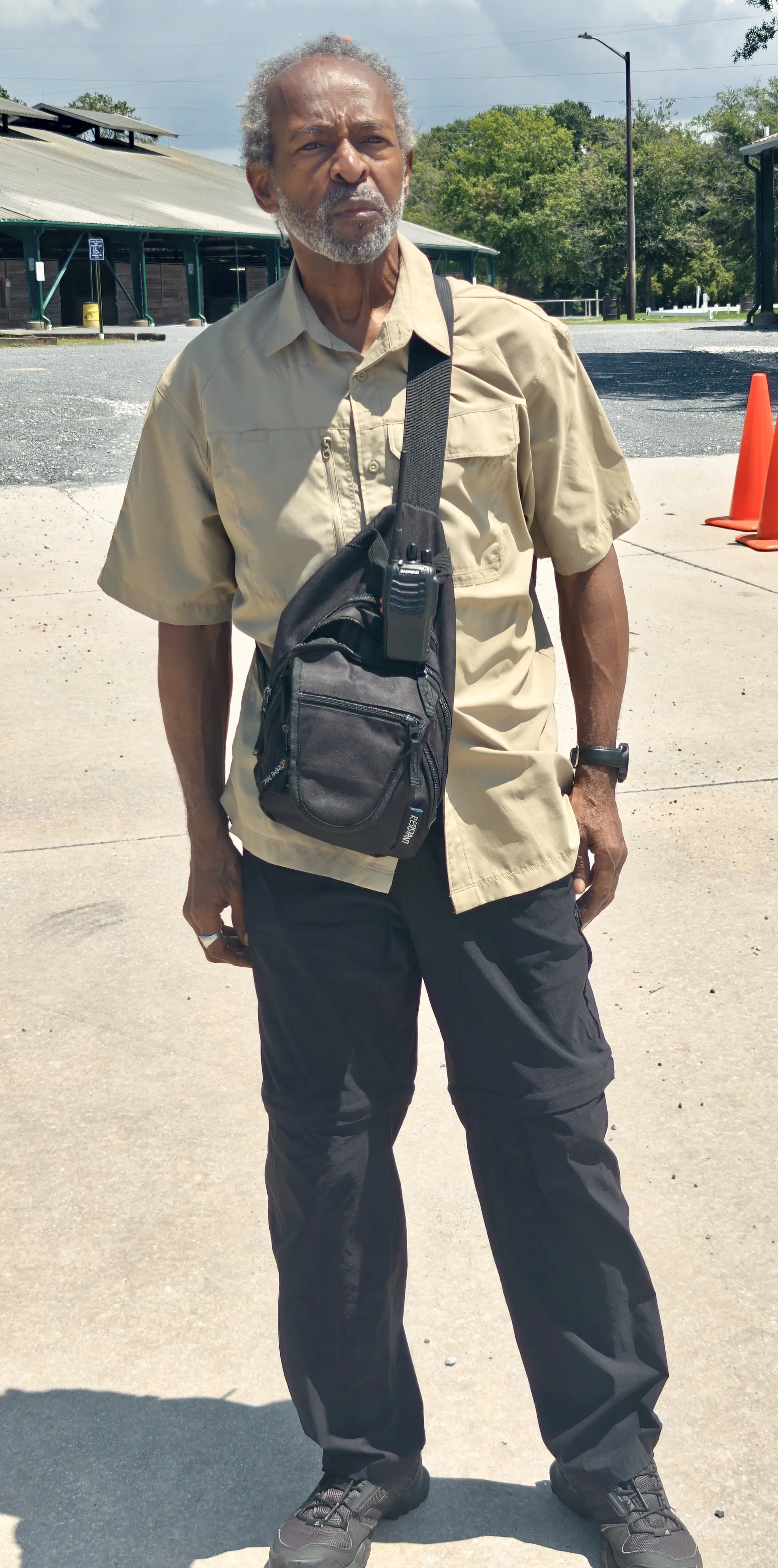 An elderly man with gray hair and beard standing outdoors, wearing a beige short-sleeve shirt, black pants, and hiking shoes. He has a small black crossbody bag with a walkie-talkie attached to it and is looking into the distance.