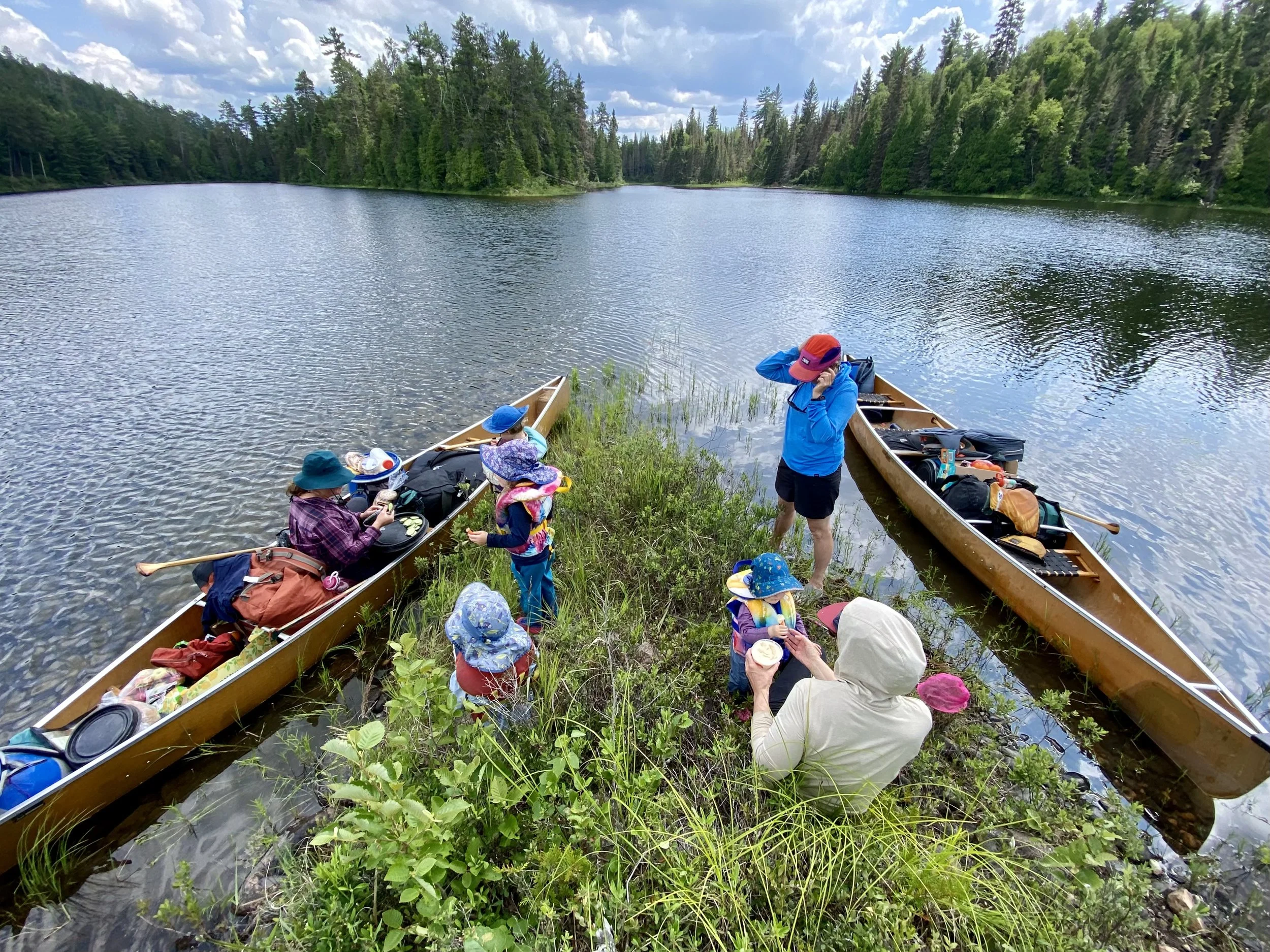 A group of people, including children and adults, preparing canoes on the shore of a lake surrounded by dense trees under a partly cloudy sky.