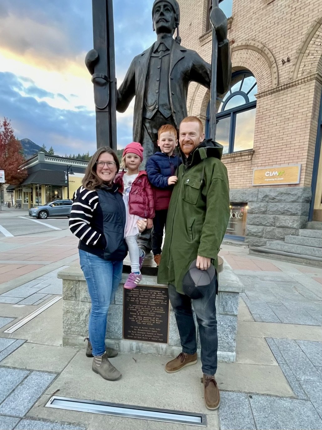 Family of four posing in front of a large bronze statue of a man holding a flag pole in a small town or city street. The family includes two children and two adults, all smiling.
