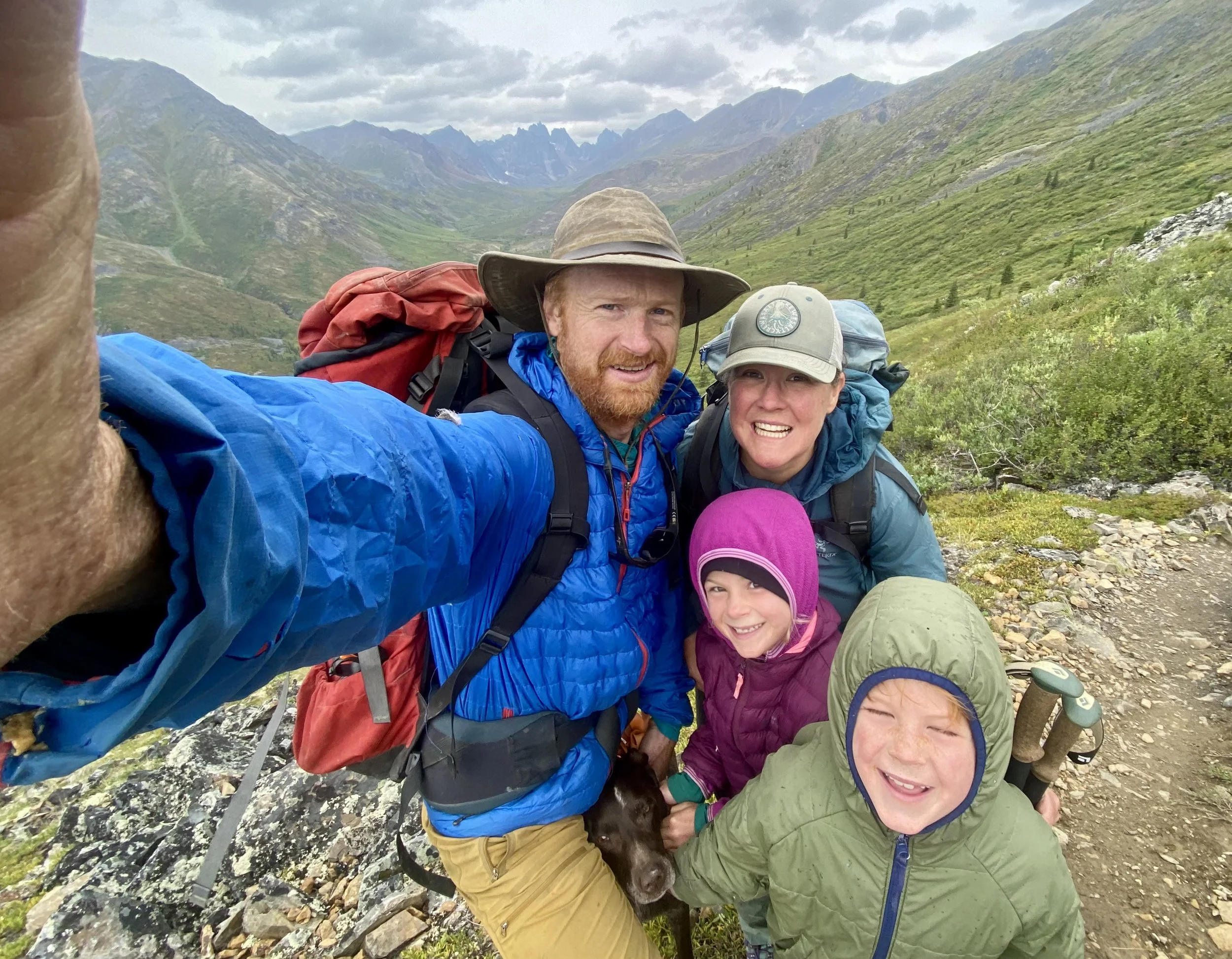 Group of hikers dressed in colorful outdoor clothing, backpacks, and hats, taking a selfie on a mountain trail with green slopes and rugged peaks in the background.