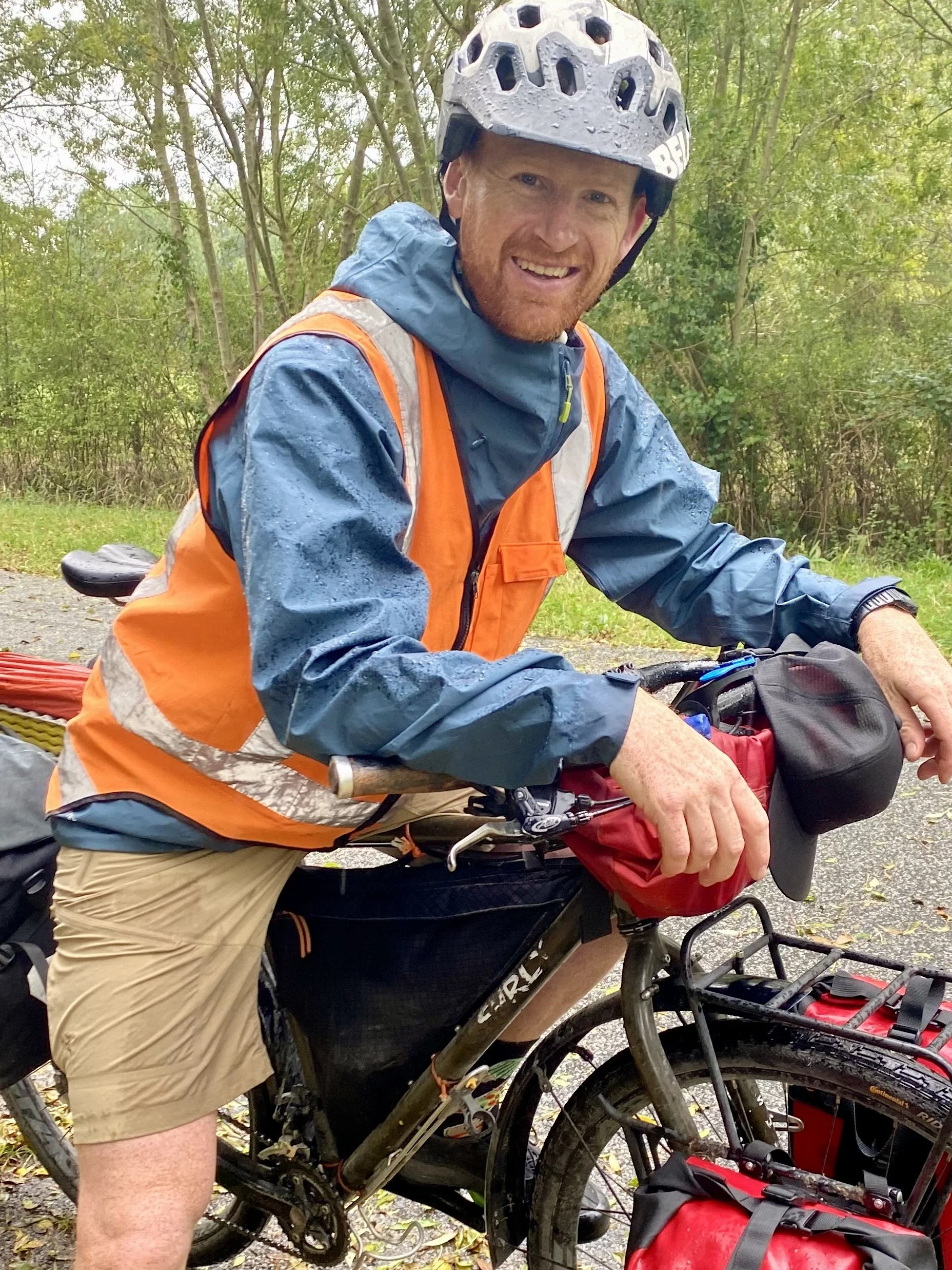 Man wearing a helmet and rain jacket sitting on a mountain bike outdoors in a wooded area, smiling at the camera.