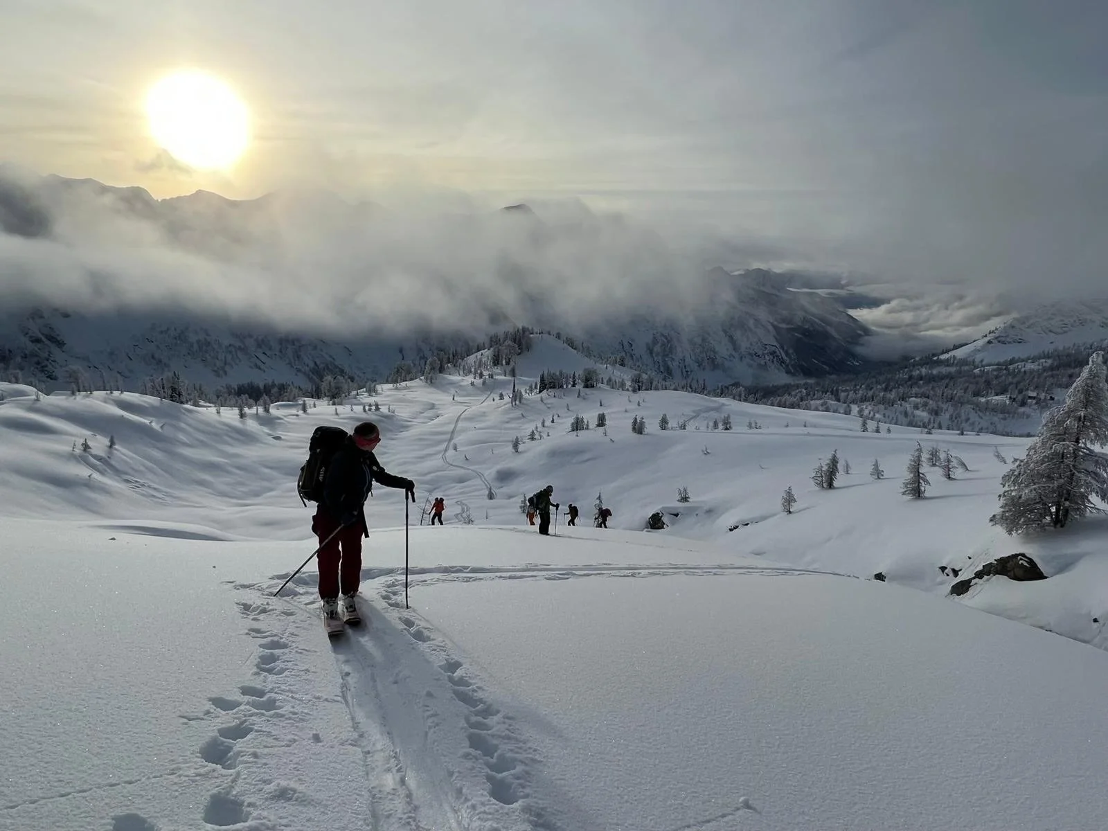 Group of people snowshoeing through snowy mountain landscape with cloudy sky and setting sun.