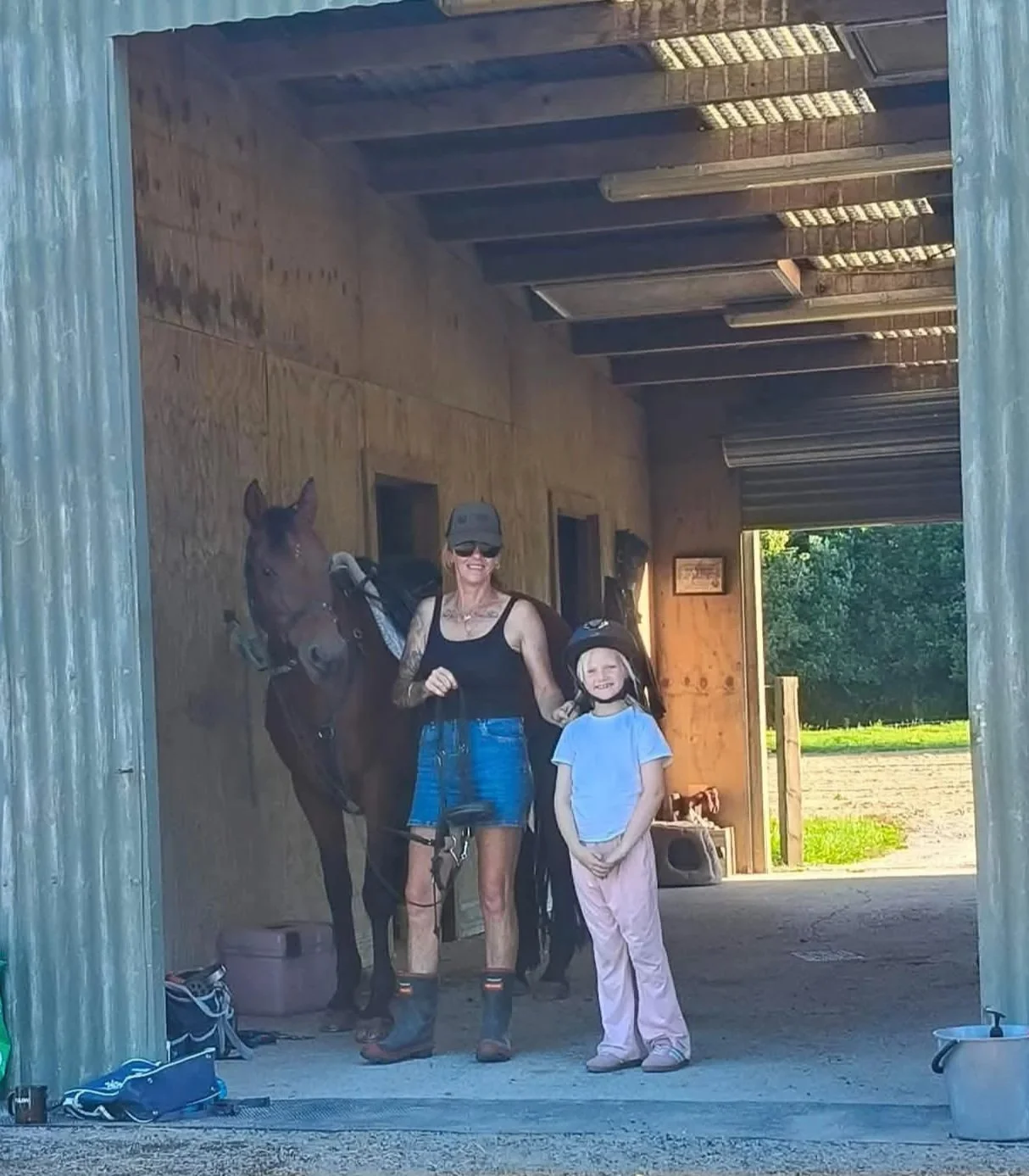A woman and a young girl standing inside a horse stable. The woman is holding the reins of a brown horse, wearing sunglasses, a hat, black tank top, and denim shorts. The girl is wearing a helmet and casual clothes, smiling. The stable has a wooden interior with bright sunlight outside.