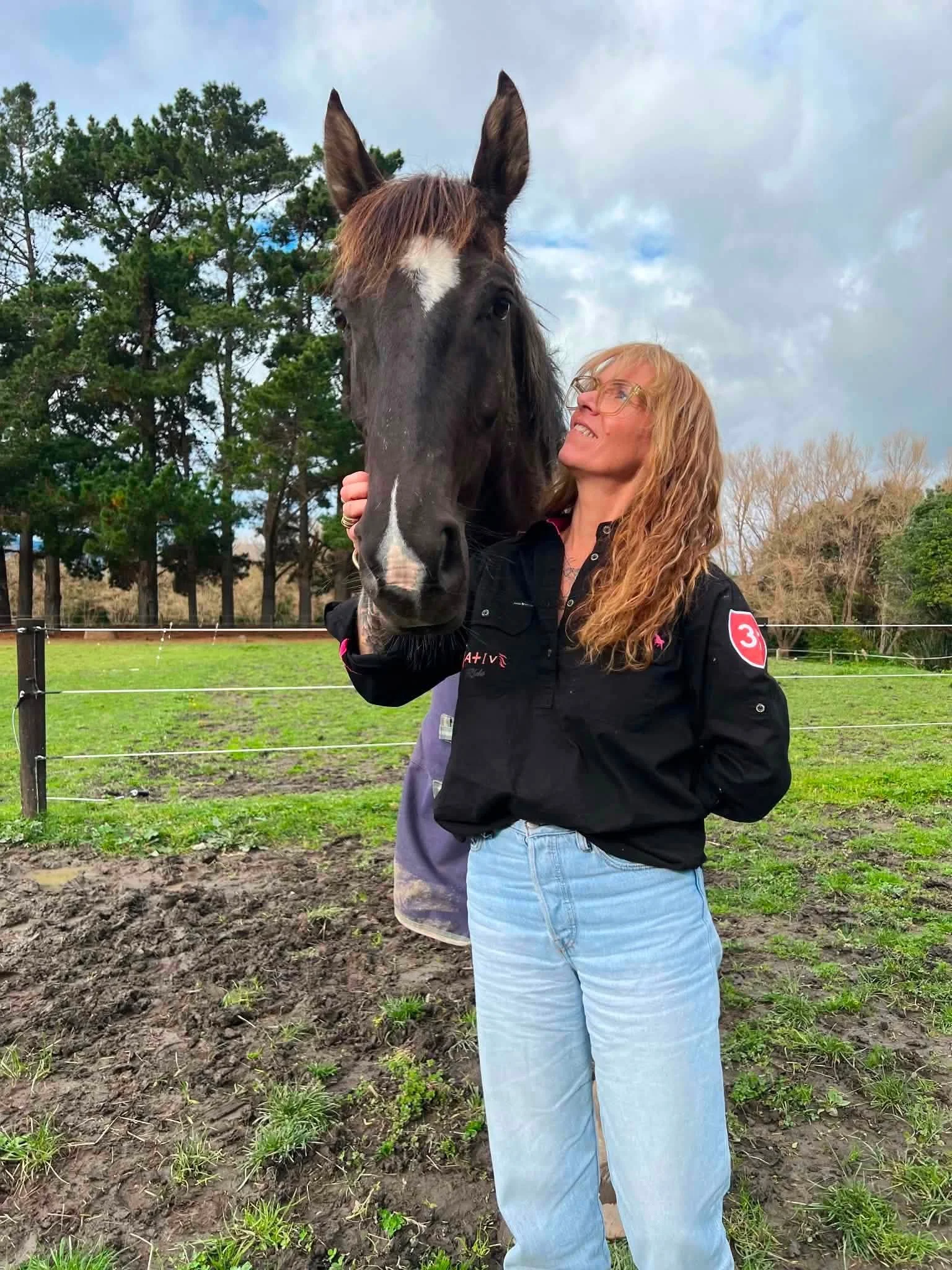 A woman with long, curly red hair and glasses stands outdoors at a farm, gently holding a black horse with a white stripe on its face. There are trees and a cloudy sky in the background.