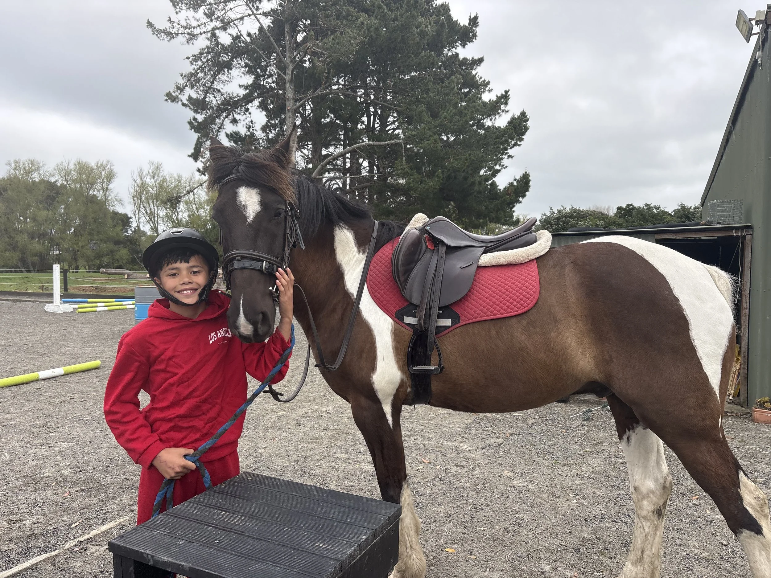A young boy wearing a red hoodie and a black helmet stands next to a brown and white horse with a black mane. The boy is smiling and touching the horse's face. The horse is saddled with a red saddle blanket and is standing on a gravel surface outdoors, with trees and a cloudy sky in the background.