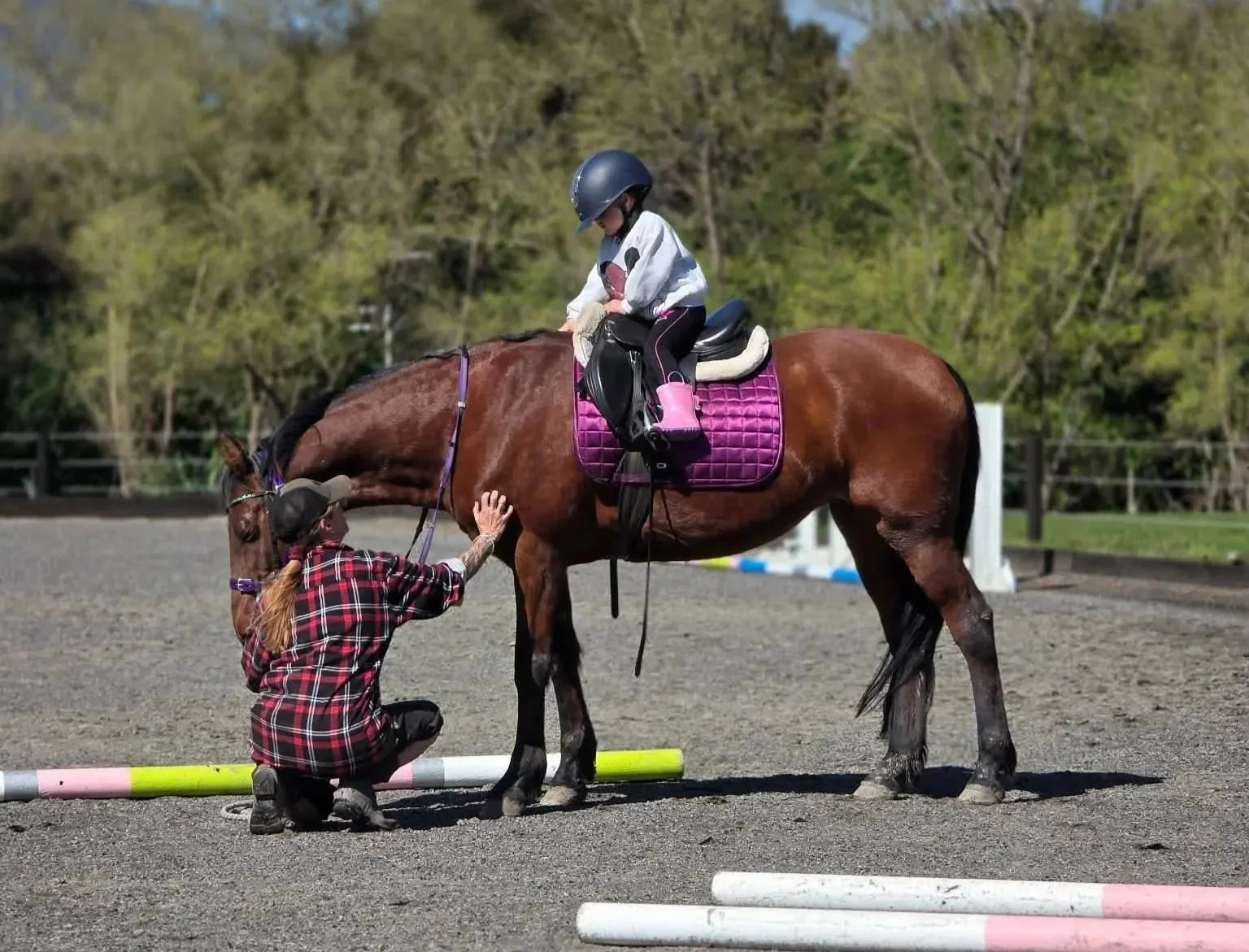 A young girl riding a brown horse with a pink saddle pad, being assisted by a woman in a plaid shirt and cap, in an outdoor riding arena with trees in the background.