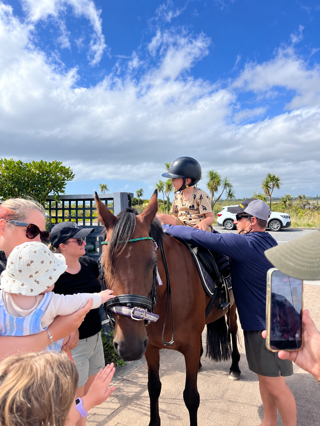 A group of people, including children and adults, gathered around a person sitting on a brown horse. The child is wearing a helmet and camouflage shirt, while the adults and other children are interacting with the rider. The scene is outdoors with blue sky, some clouds, and palm trees in the background.