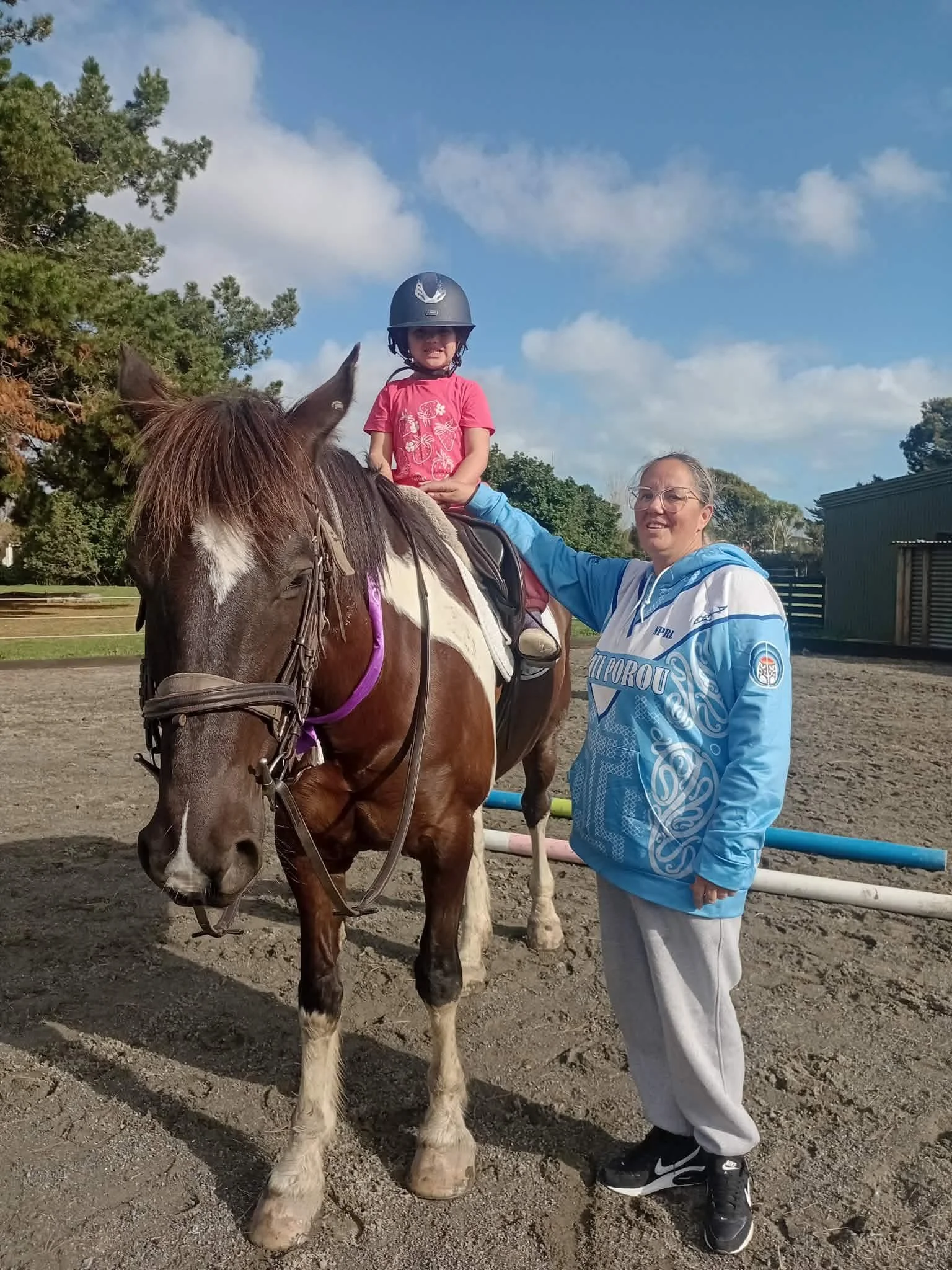 A young girl wearing a black helmet and a pink t-shirt sits on a white and brown horse. An older woman in glasses, a blue and white sports jacket, and gray pants stands next to them, holding the horse's reins and smiling. The background features trees, a blue sky with clouds, and a wooden structure.