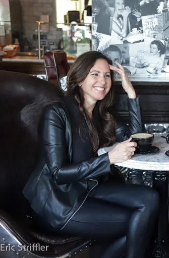 A woman coach with long brown hair, wearing a cool black leather jacket, sitting in a cafe, smiling, holding a black coffee cup, talking with a client.