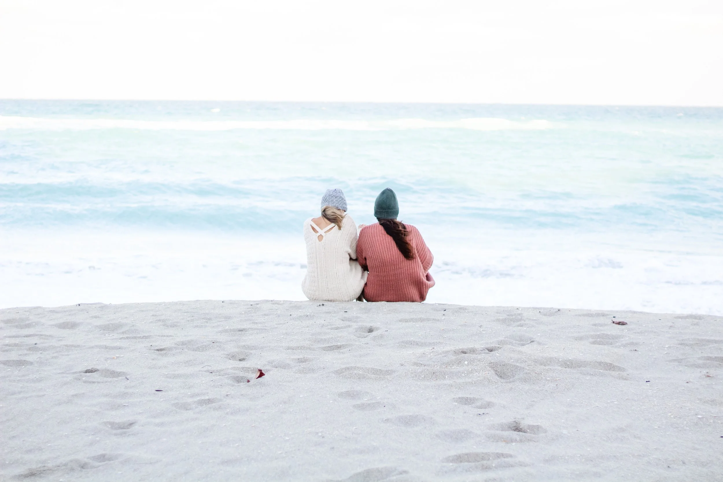 Two women sitting on the sandy beach facing the ocean and just holding space for one another.