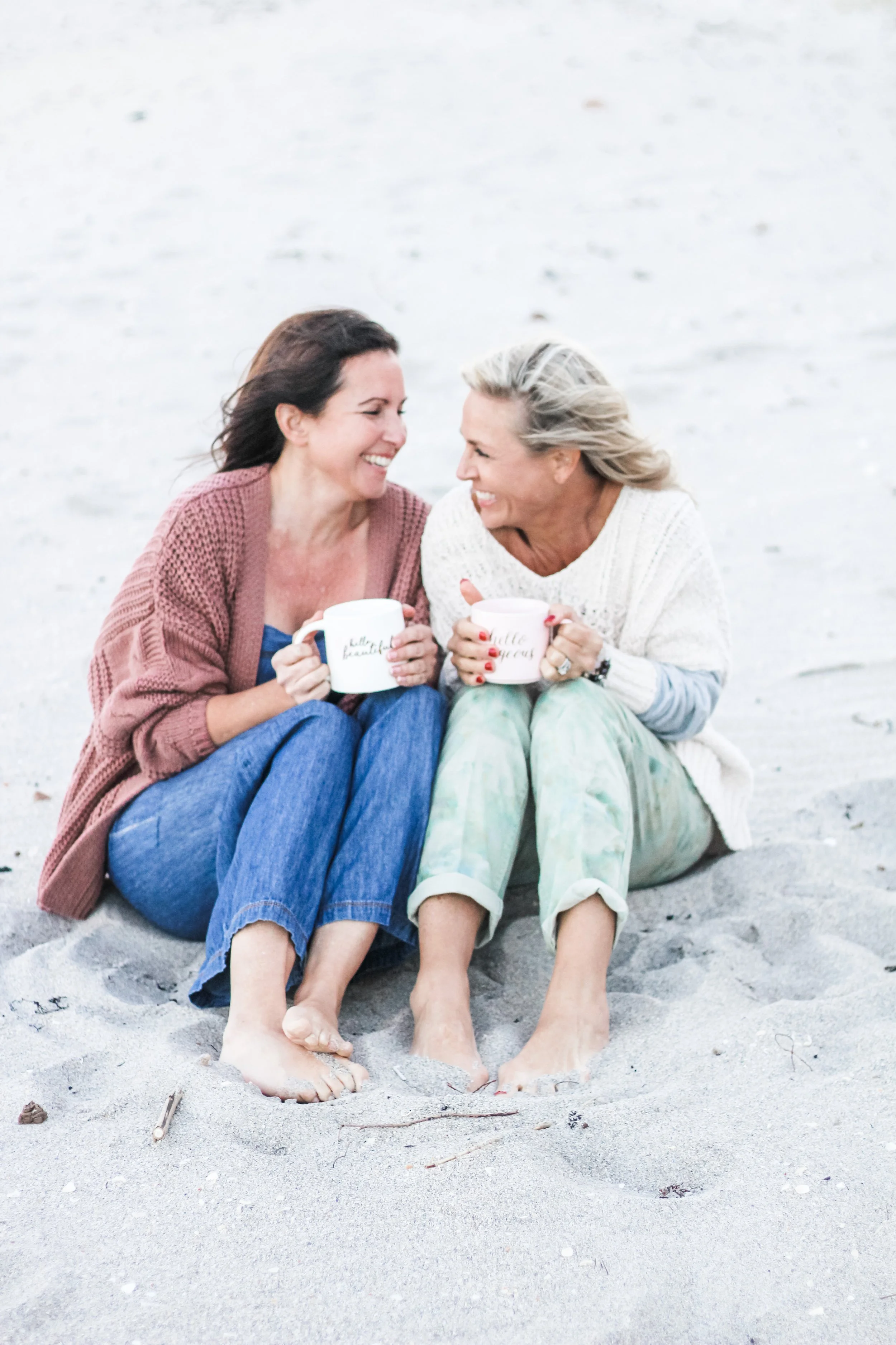 Two women sitting close together on a sandy beach, smiling and holding white coffee mugs, laughing and supporting each other.