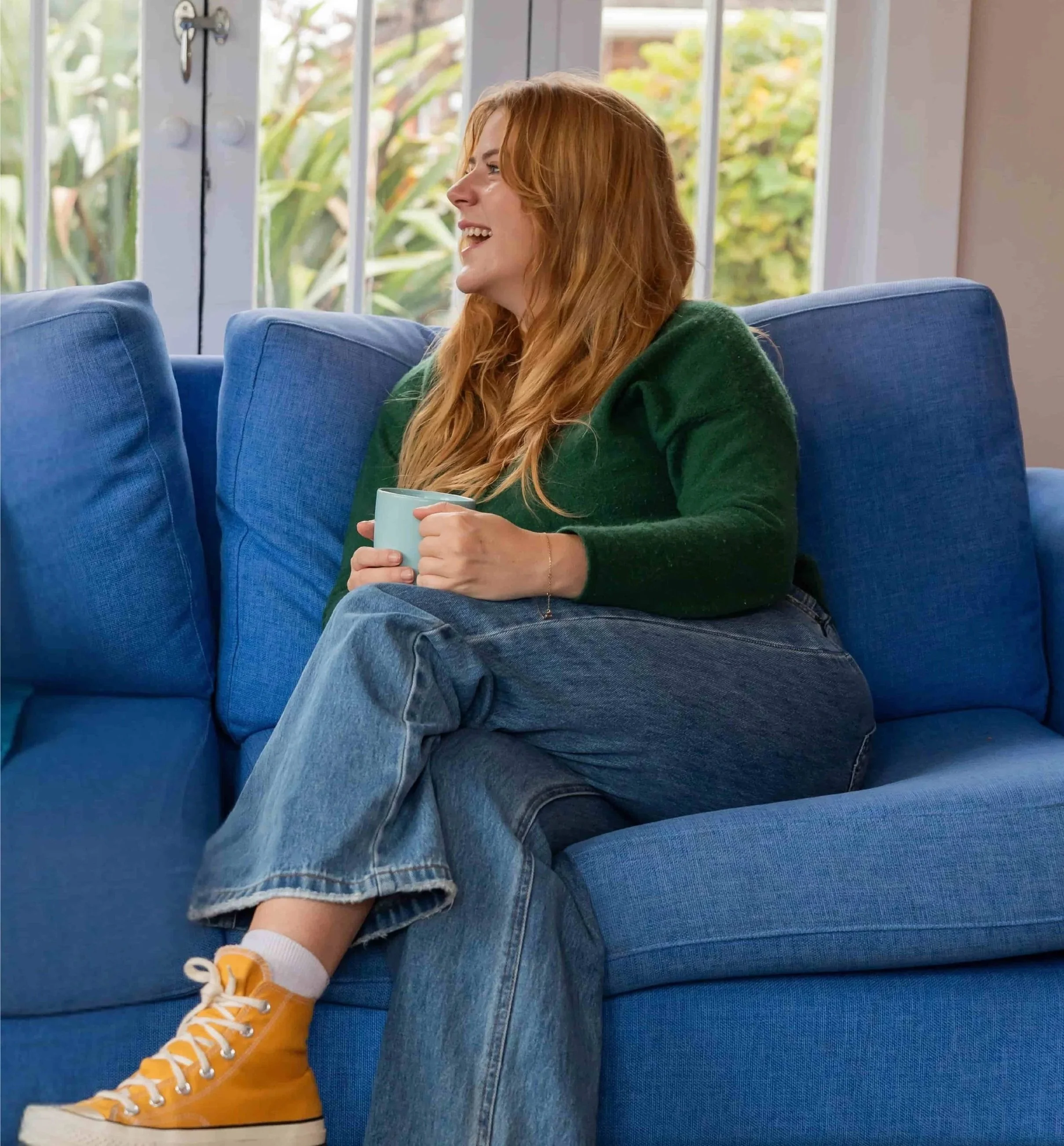 A woman with red hair, wearing a green sweater, blue jeans, and yellow sneakers sits on a blue couch, holding a light blue mug. She is smiling and looking to her right in a room with large windows showing green foliage outside.