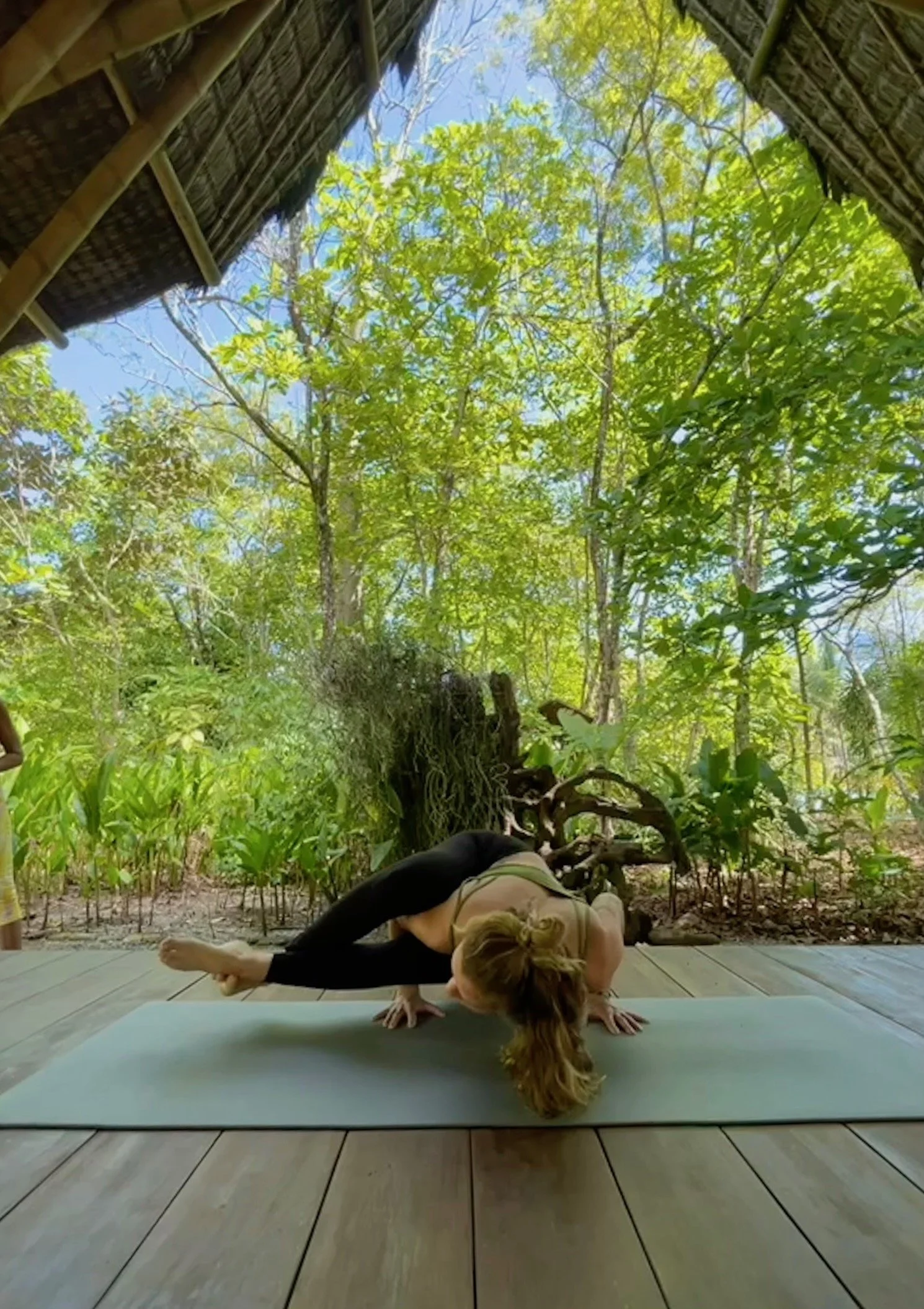 Woman practicing yoga on a mat beneath a thatched hut surrounded by lush tropical forest
