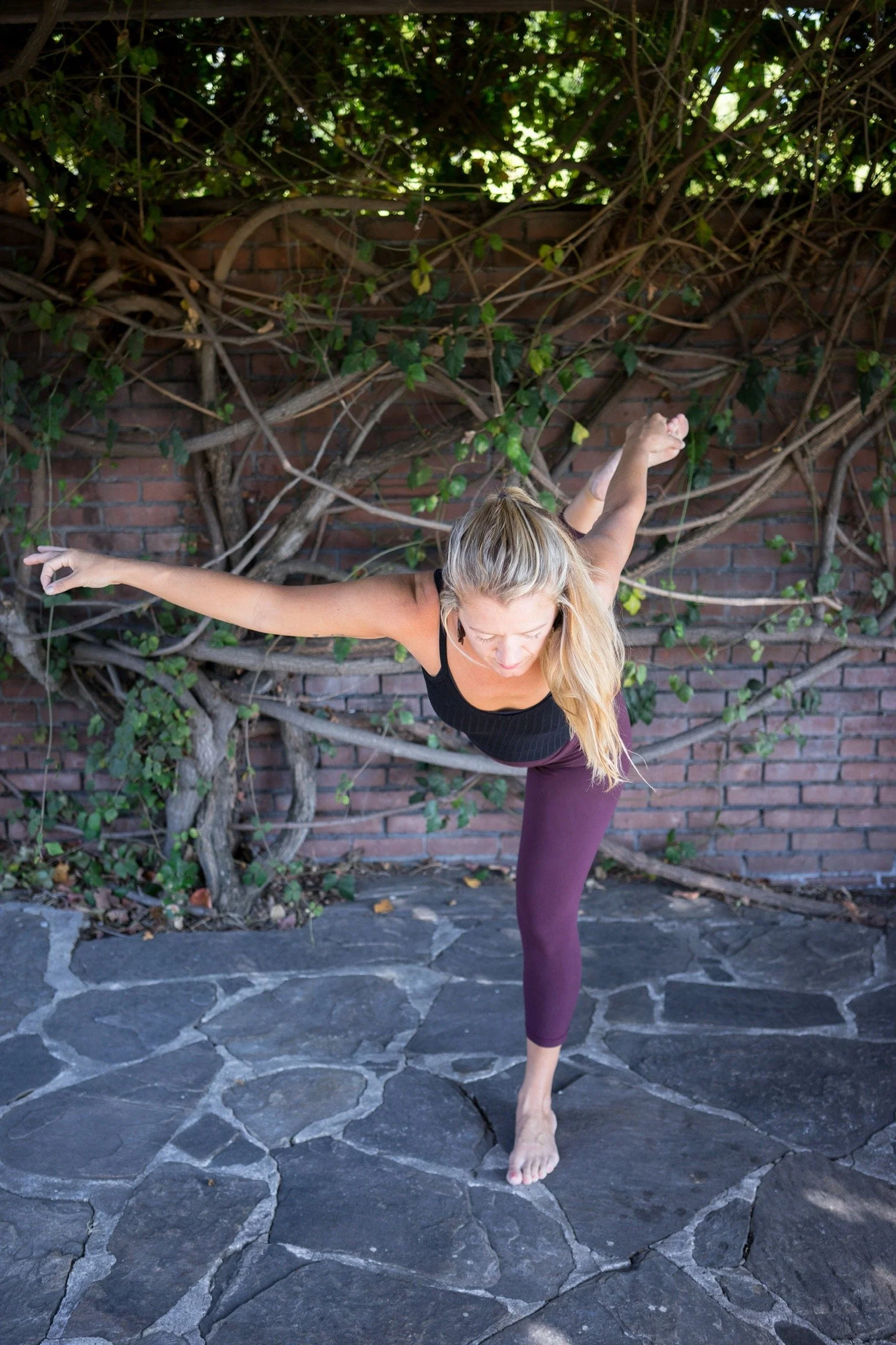 Alyson LeBeau practicing a yoga balance pose barefoot on a stone patio in front of an ivy-covered brick wall