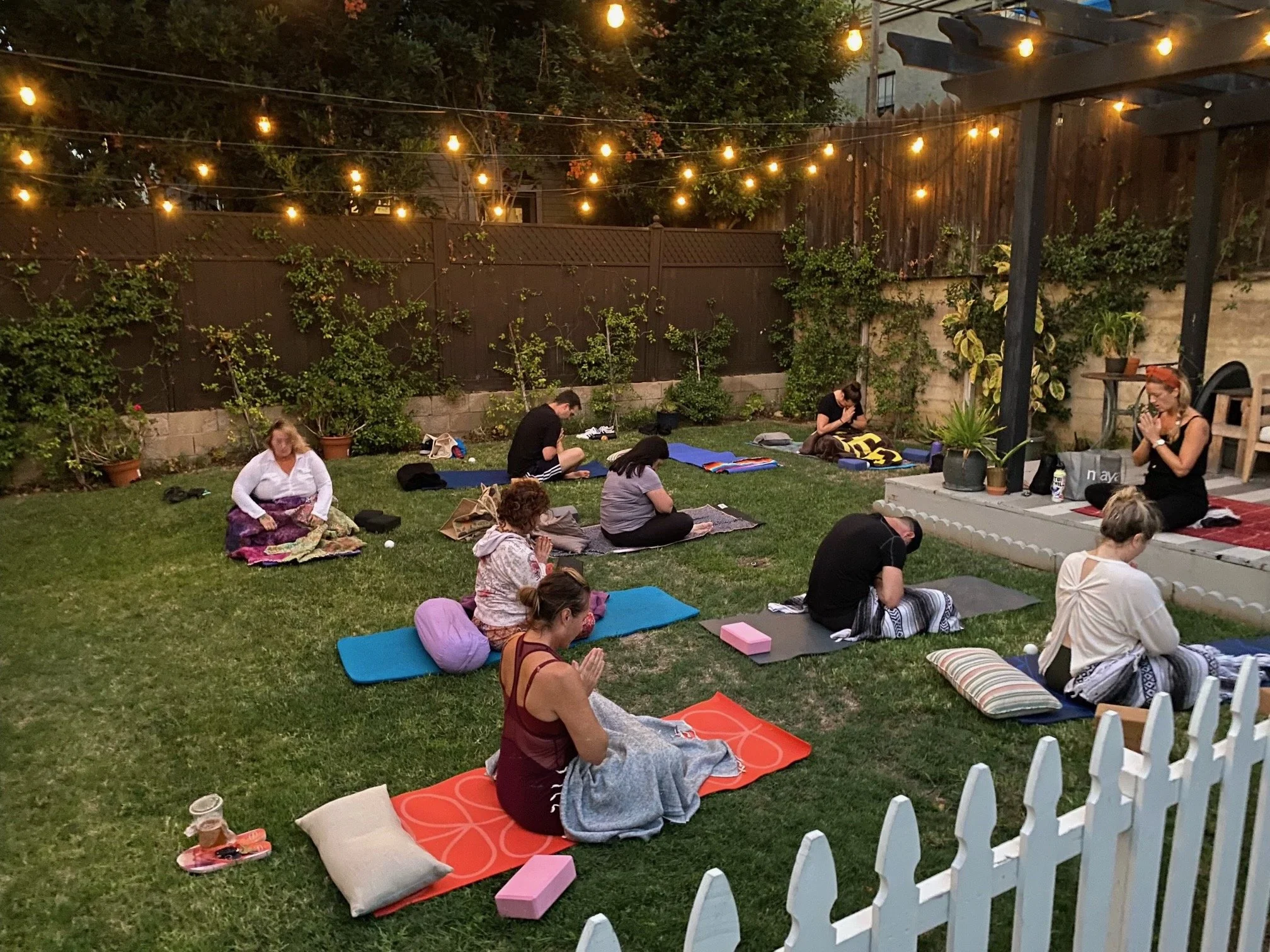 Outdoor evening yoga class with people on mats in a grassy backyard decorated with string lights