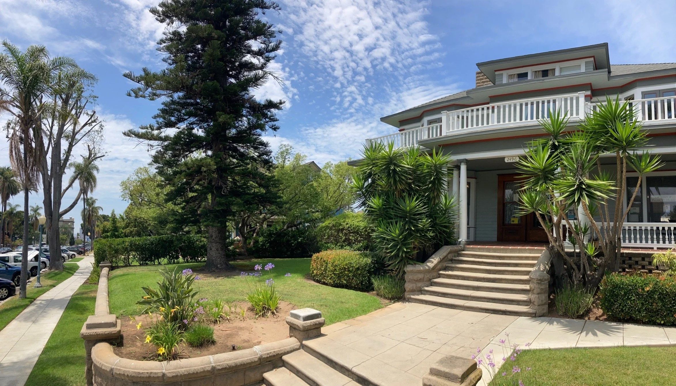 Historic gray house with white balconies, landscaped front garden, palm trees, and sidewalk under a blue sky in San Diego