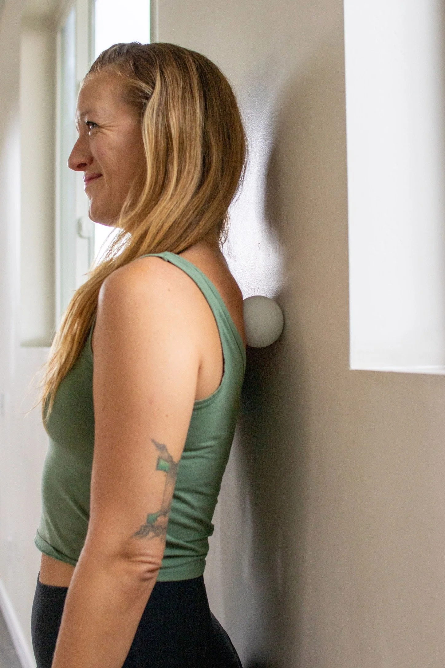 Side profile of a smiling woman with long blonde hair wearing a green tank top indoors using a lacrosse ball for myofascial release