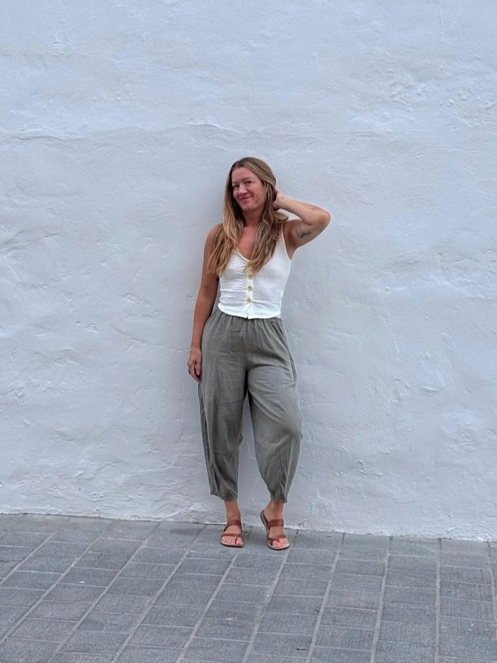 Alyson LeBeau posing in a white button-front top, loose gray pants, and sandals against a white stucco wall.
