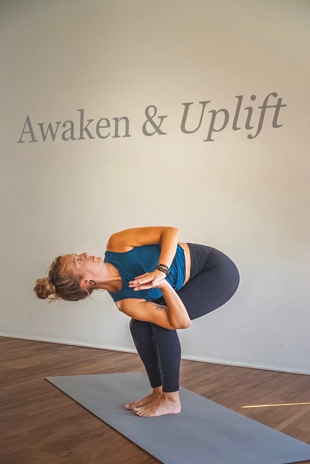 Woman practicing yoga in a twisted squat pose on a mat beneath “Awaken & Uplift” wall lettering